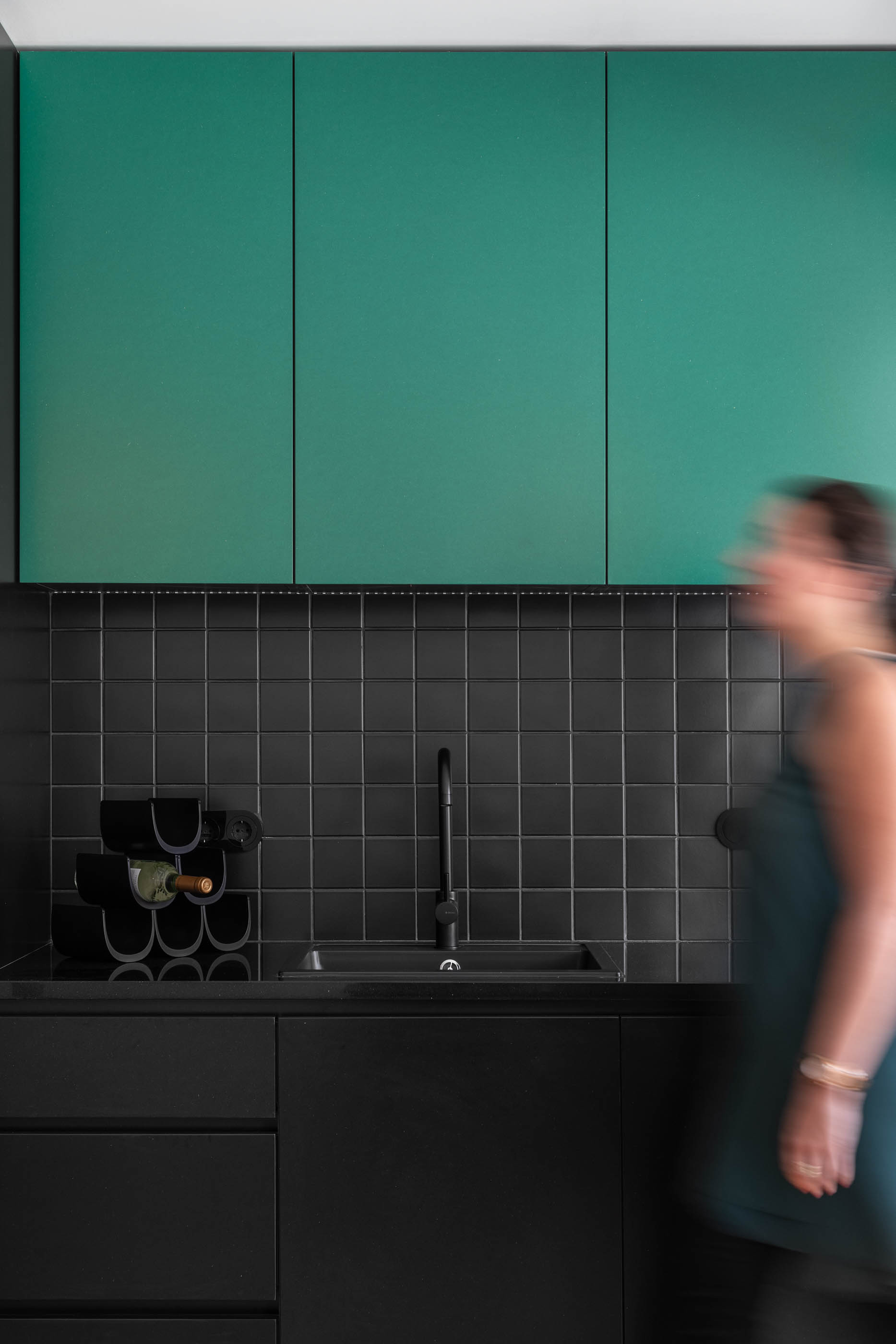 Cozinha com armários verdes e pretos, azulejos pretos e mulher a passar. — Apartamento Morro arquitetura Hinterland Architecture Studio em Vila Nova de Gaia
