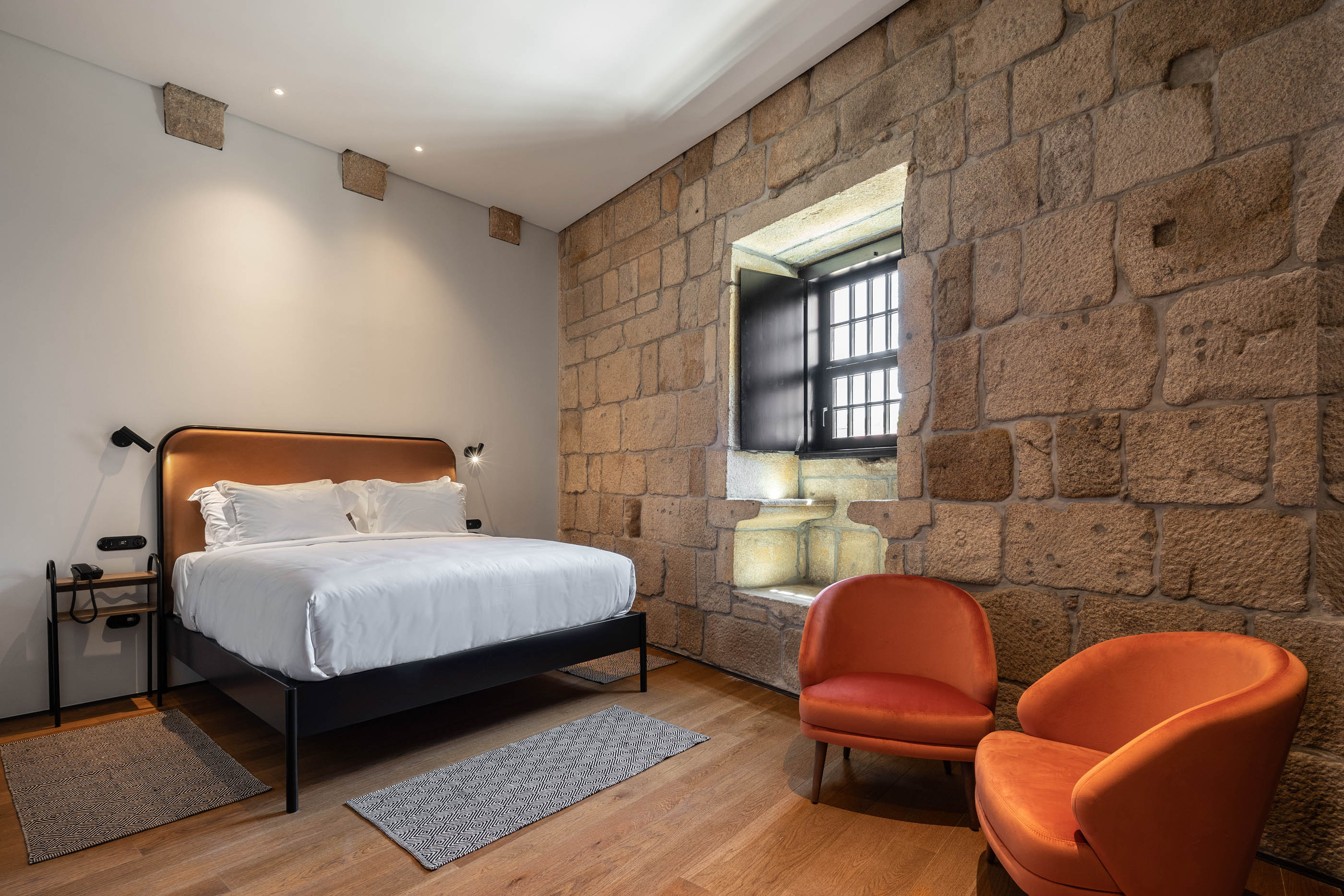 Quarto com parede em pedra original, cama de casal e cadeirões laranjas, estilo rústico moderno. — Exmo Hotel arquitetura Floret Arquitectura em Porto