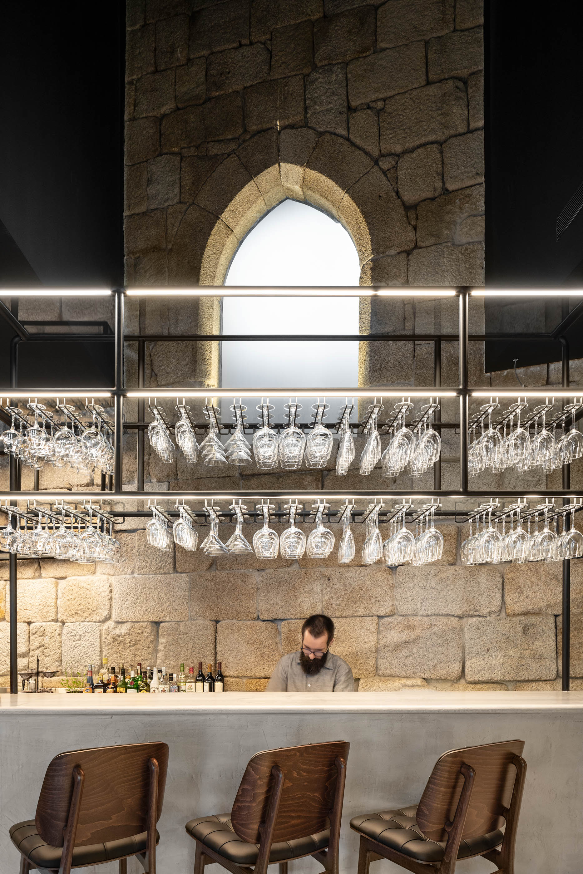 Balcão de bar com paredes em pedra original e prateleiras com copos, funcionário atrás do balcão. — Exmo Hotel arquitetura Floret Arquitectura em Porto