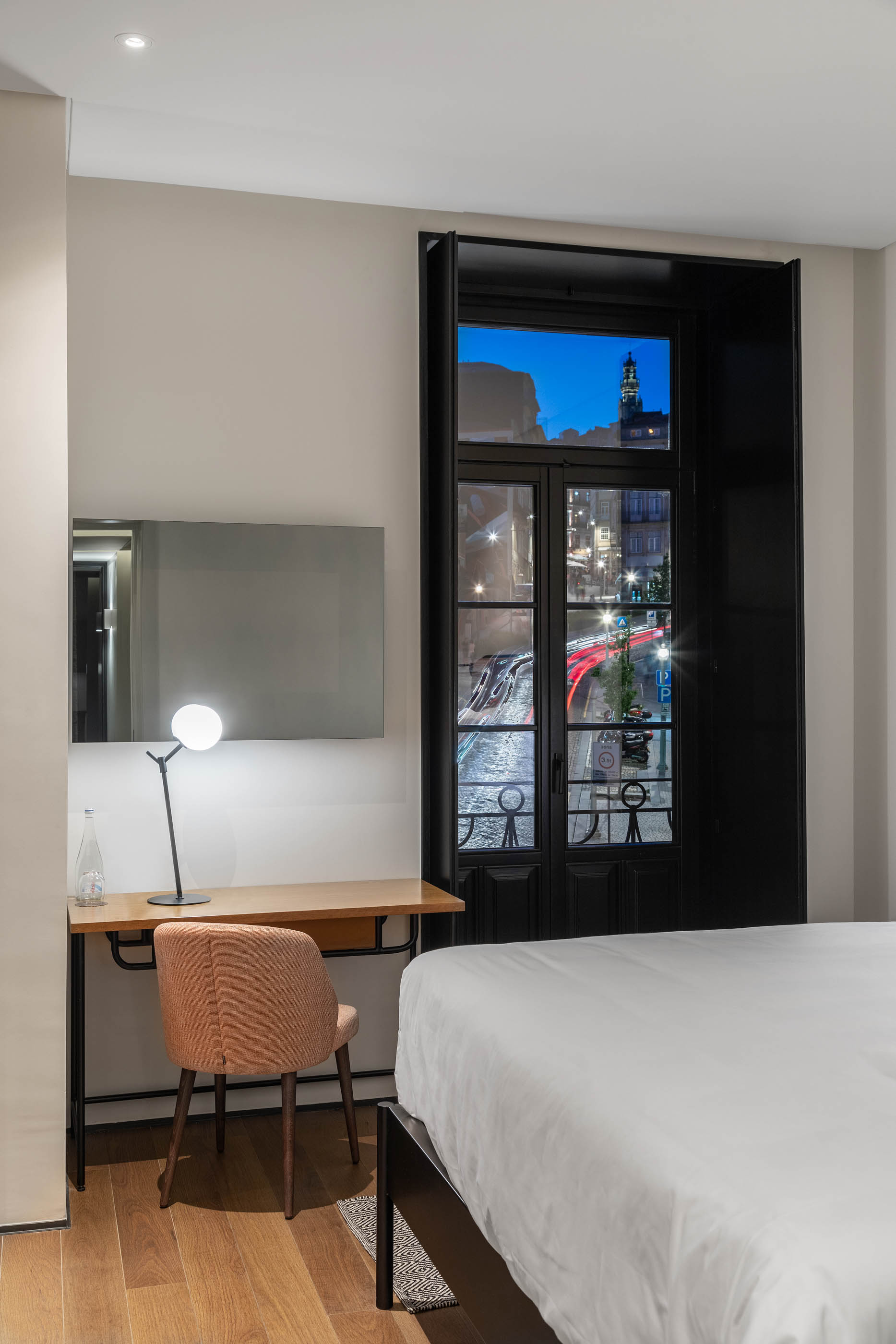 Quarto de hotel com secretária em madeira, candeeiro moderno e vista noturna da cidade através da janela. — Exmo Hotel arquitetura Floret Arquitectura em Porto