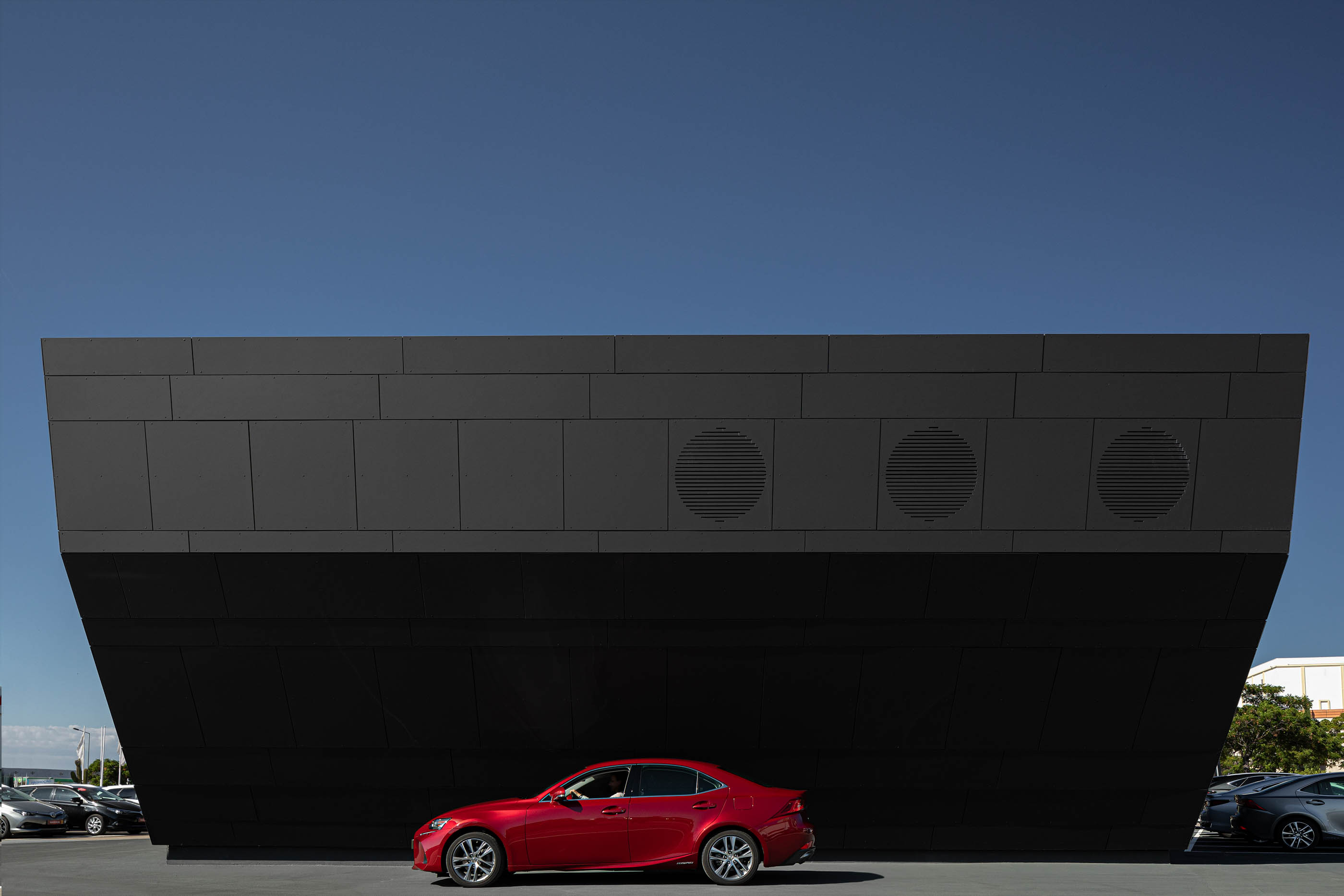 Fachada lateral do stand Lexus, com revestimento escuro, detalhes circulares e um carro vermelho estacionado em frente. — Stand Lexus Faro arquitetura Rarcon Arquitectura em Portugal