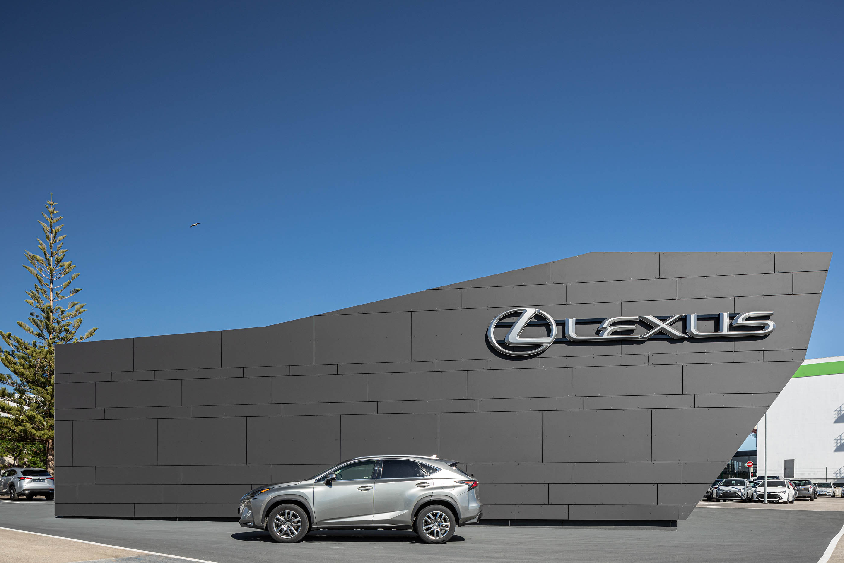 Fachada lateral do stand Lexus, com revestimento cinzento, logótipo prateado e um carro cinzento estacionado em frente. — Stand Lexus Faro arquitetura Rarcon Arquitectura em Portugal