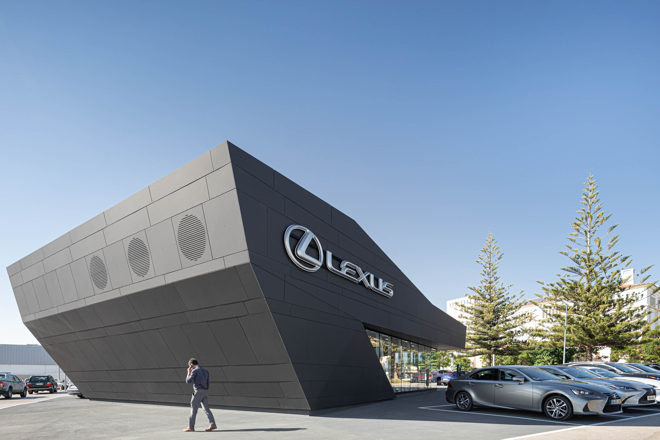 Perspectiva angular do stand Lexus, com revestimento cinzento, logótipo prateado e carros estacionados em frente. — Stand Lexus Faro arquitetura Rarcon Arquitectura em Portugal