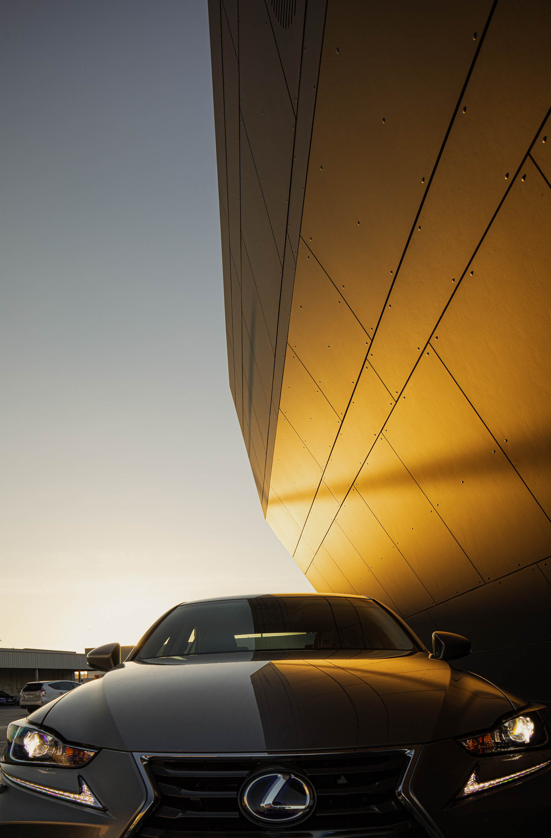 Detalhe da fachada do stand Lexus ao pôr do sol, com revestimento escuro e reflexos dourados. — Stand Lexus Faro arquitetura Rarcon Arquitectura em Portugal