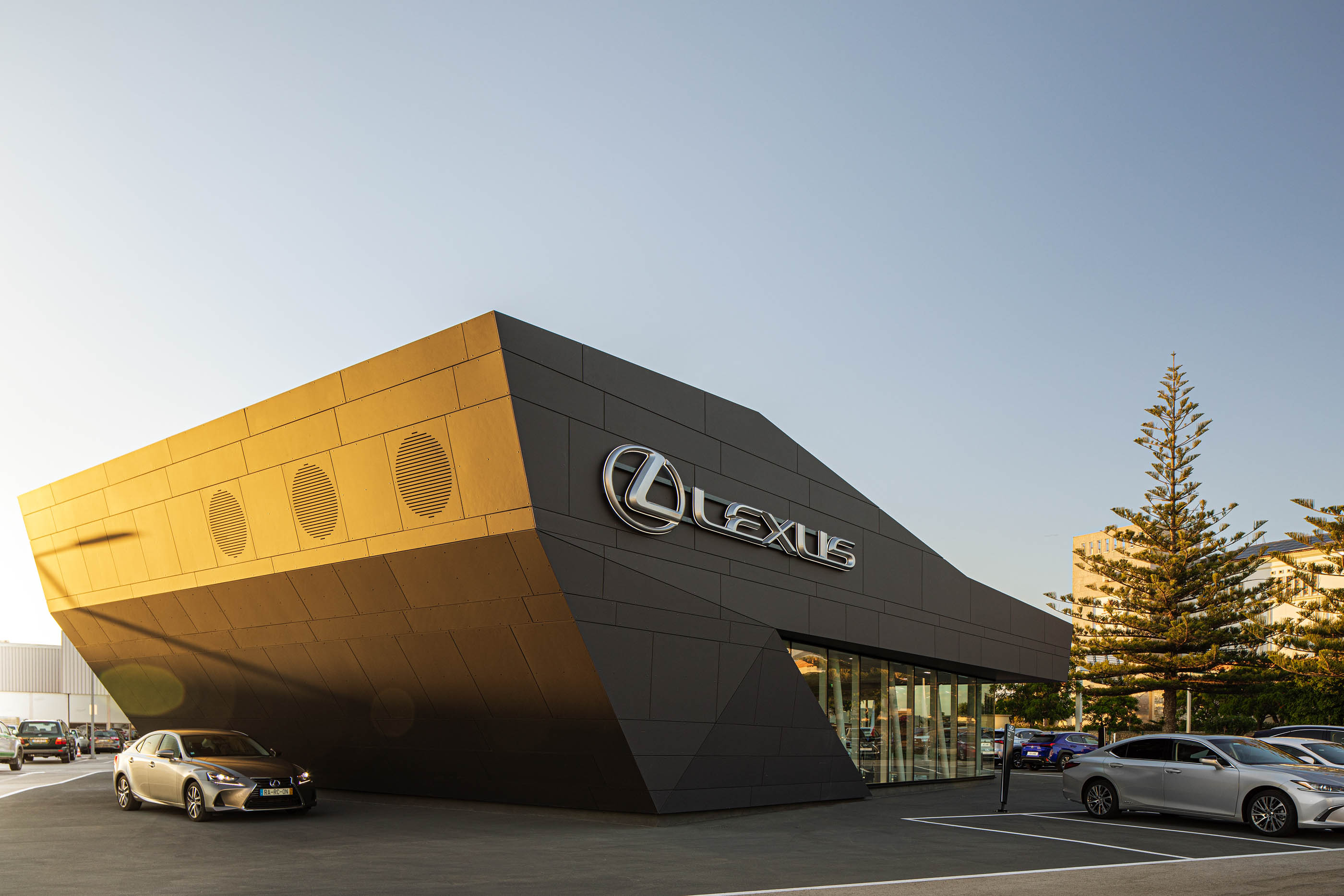 Vista frontal do stand Lexus, com revestimento cinzento, logótipo prateado e carros estacionados em frente. — Stand Lexus Faro arquitetura Rarcon Arquitectura em Portugal