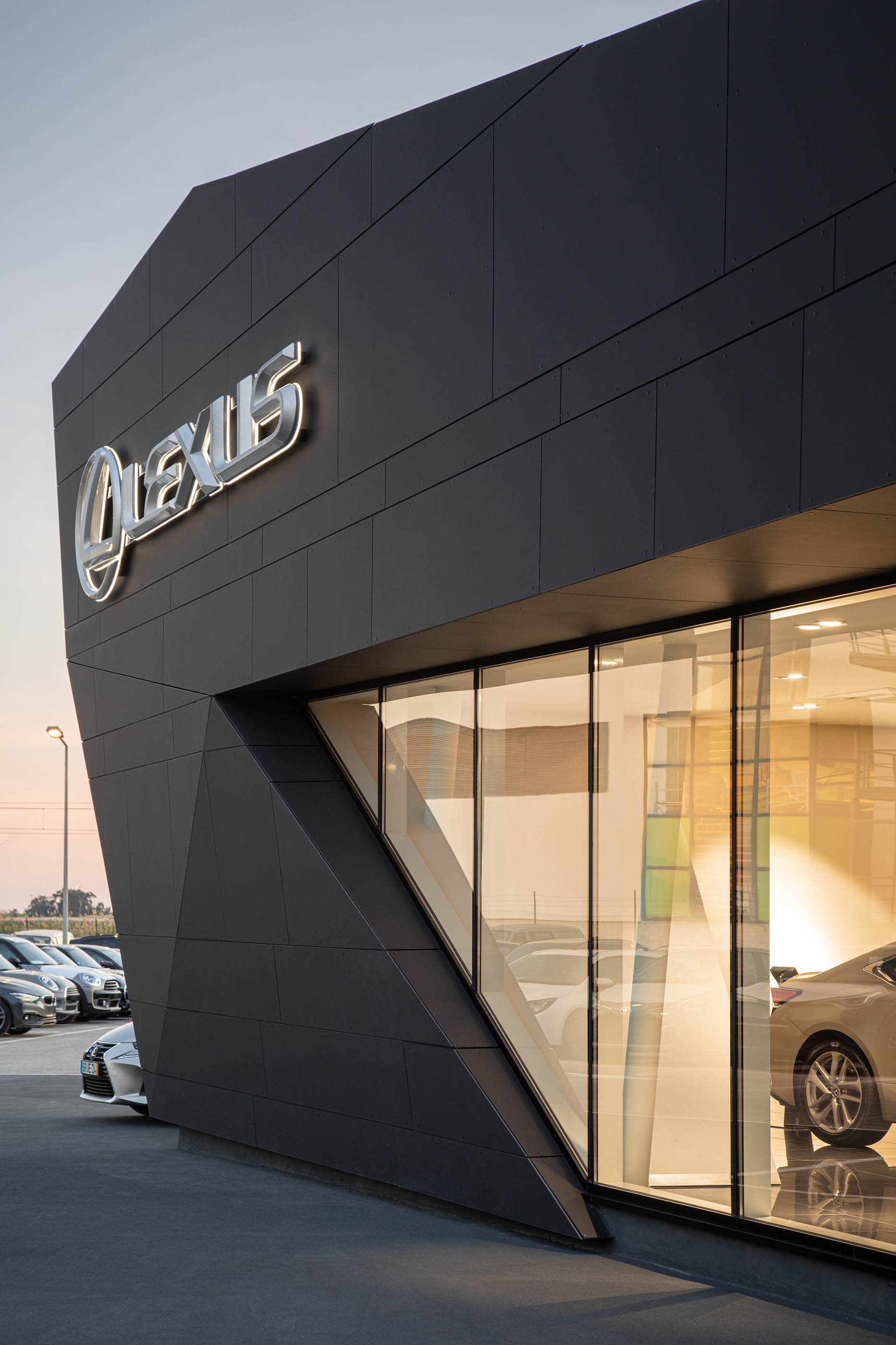 Detalhe da fachada do stand Lexus, com revestimento escuro, janelas triangulares e carros estacionados em frente. — Stand Lexus Faro arquitetura Rarcon Arquitectura em Portugal