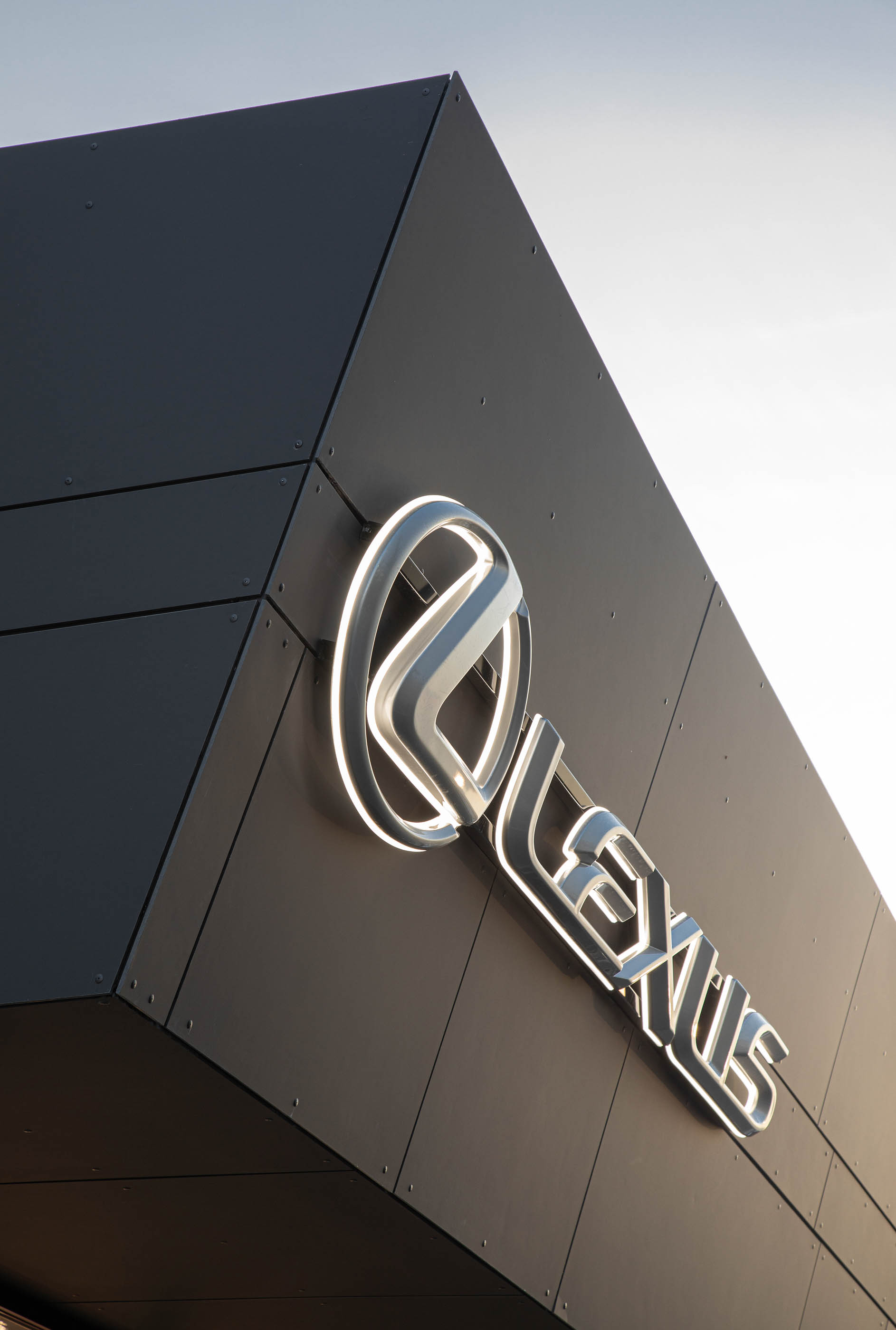 Close-up do logótipo Lexus na fachada do stand, com revestimento escuro e iluminação suave. — Stand Lexus Faro arquitetura Rarcon Arquitectura em Portugal