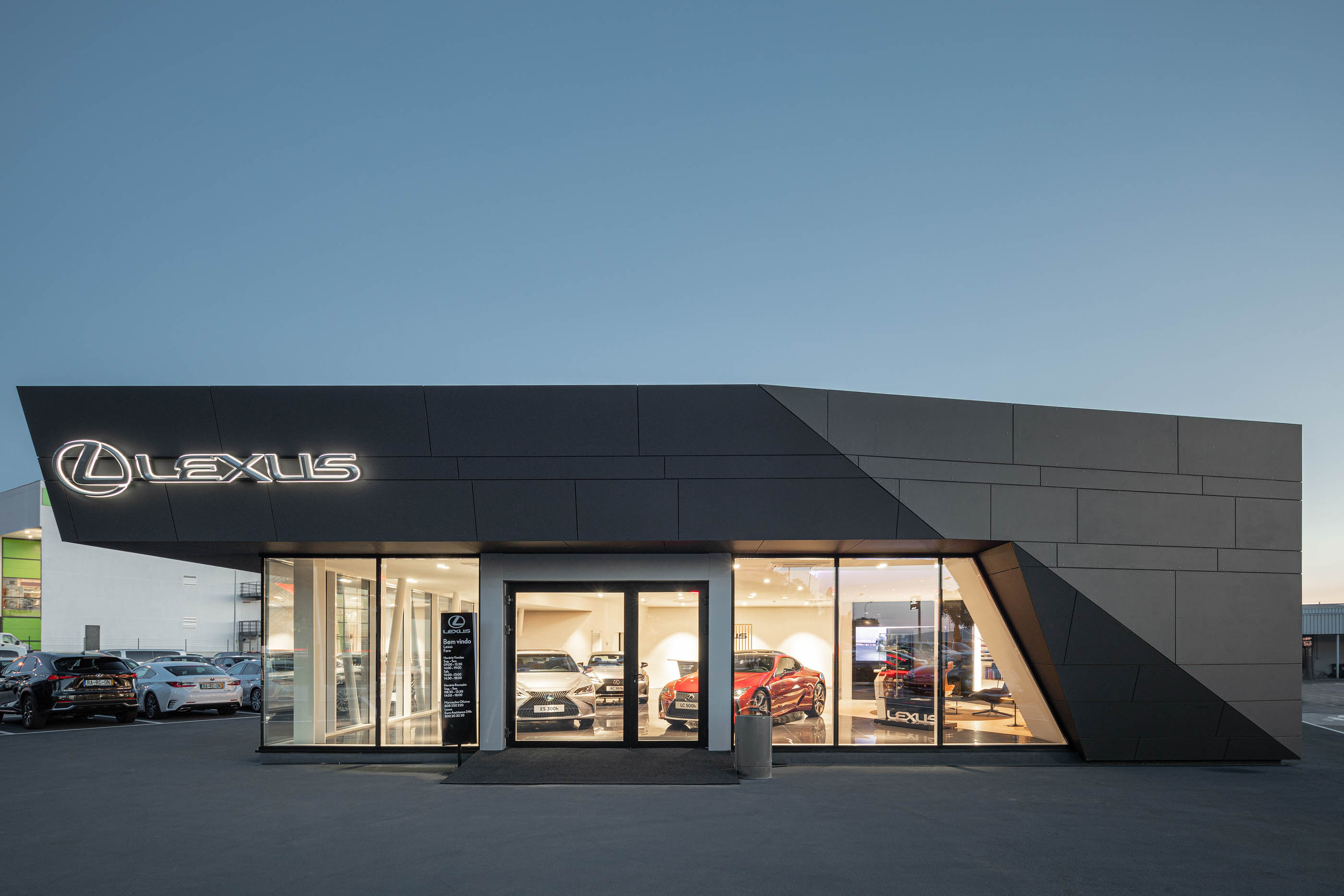 Vista noturna do stand Lexus, com revestimento cinzento, logótipo iluminado e carros expostos nas janelas. — Stand Lexus Faro arquitetura Rarcon Arquitectura em Portugal