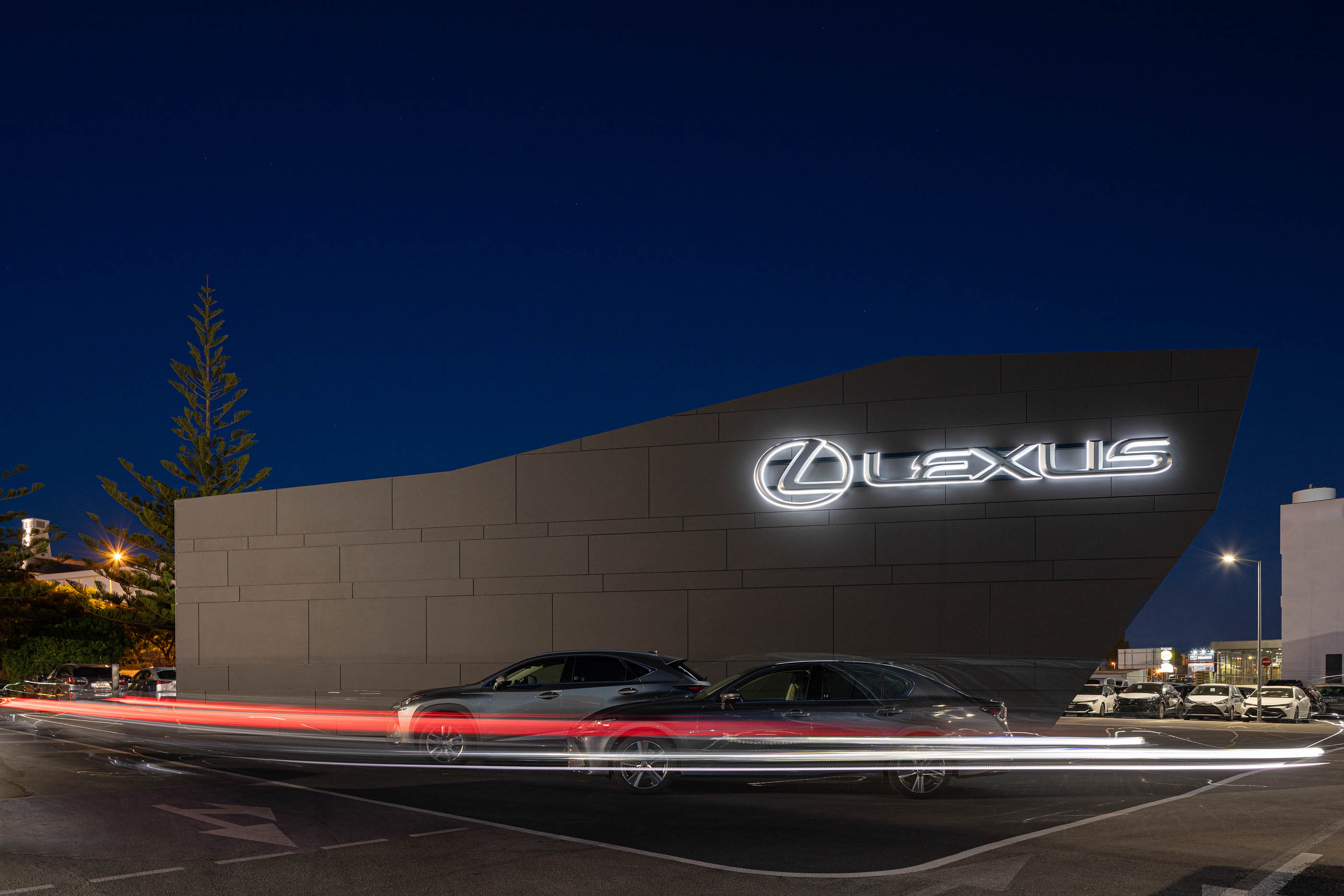 Vista noturna do stand Lexus, com revestimento cinzento, logótipo iluminado e rastos de luz dos carros em movimento. — Stand Lexus Faro arquitetura Rarcon Arquitectura em Portugal