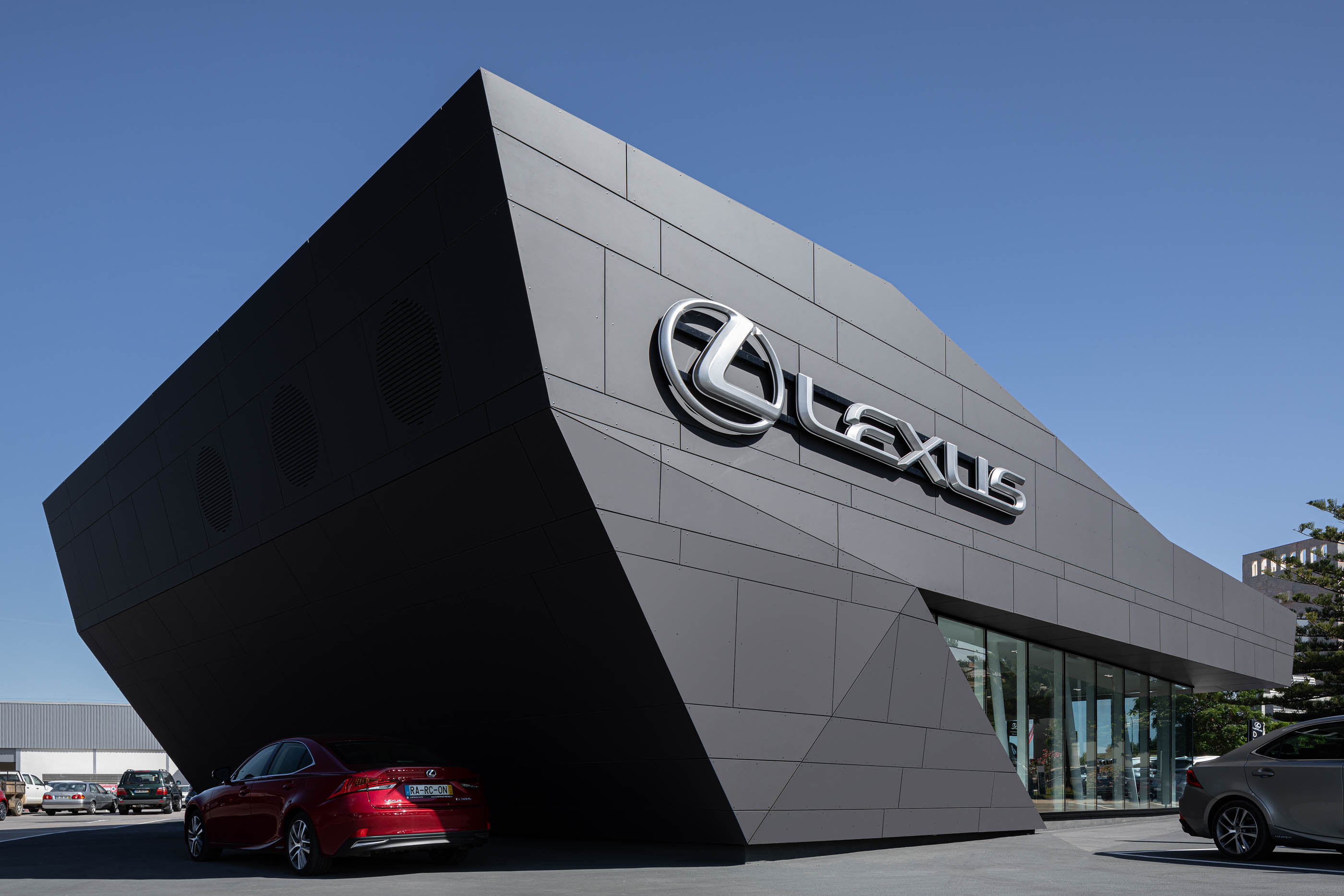 Edifício Lexus com design angular e revestimento escuro, destacando o logótipo prateado e carros estacionados em frente. — Stand Lexus Faro arquitetura Rarcon Arquitectura em Portugal