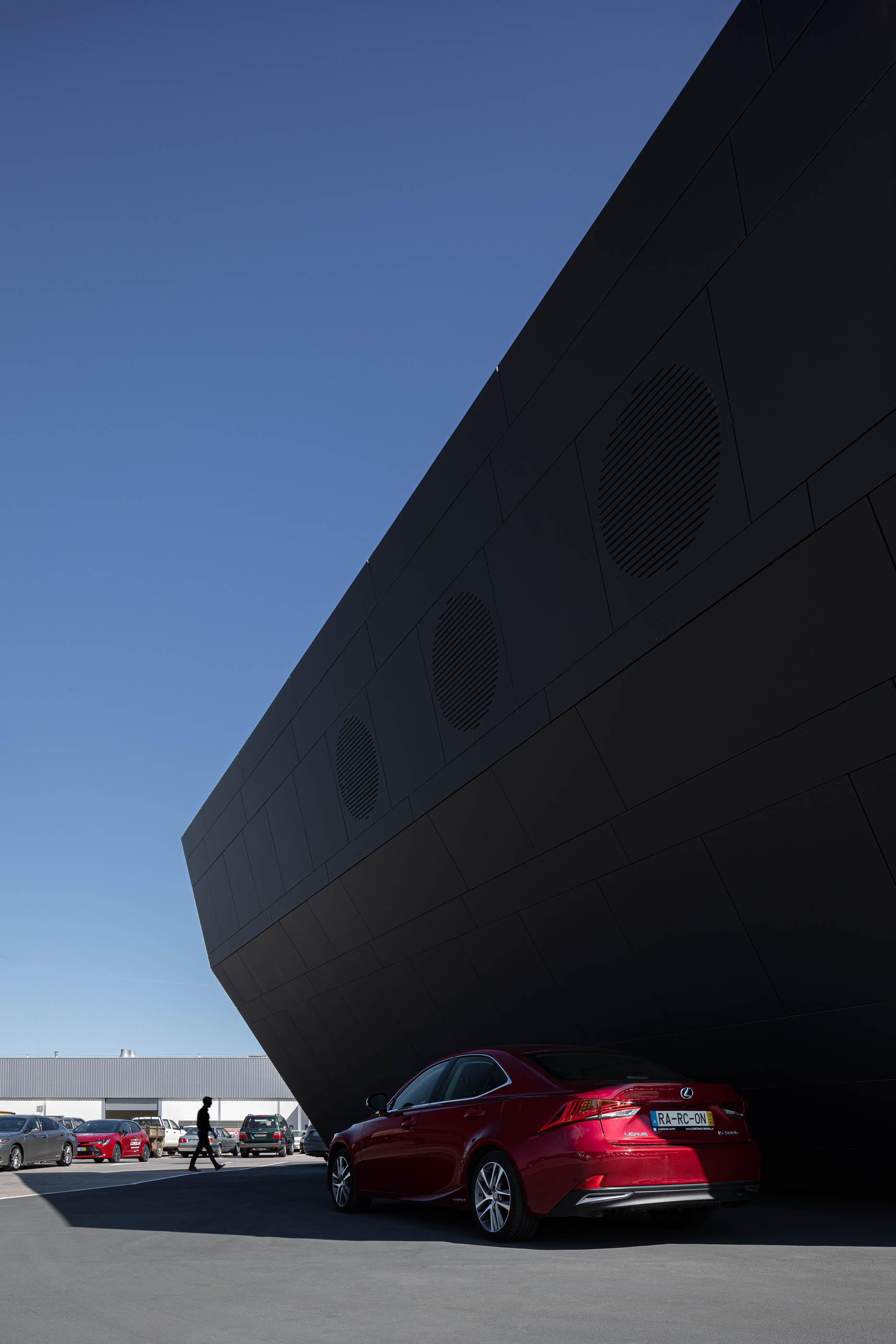 Vista lateral do edifício Lexus, com revestimento escuro, detalhes circulares e um carro vermelho estacionado em primeiro plano. — Stand Lexus Faro arquitetura Rarcon Arquitectura em Portugal