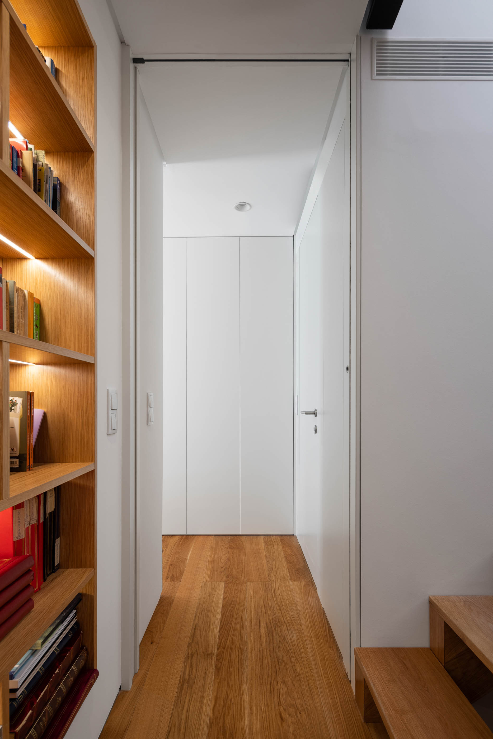 Corredor com estante de madeira iluminada e porta branca ao fundo. — Casa GD arquitetura Esquissos Arquitectura em Portugal