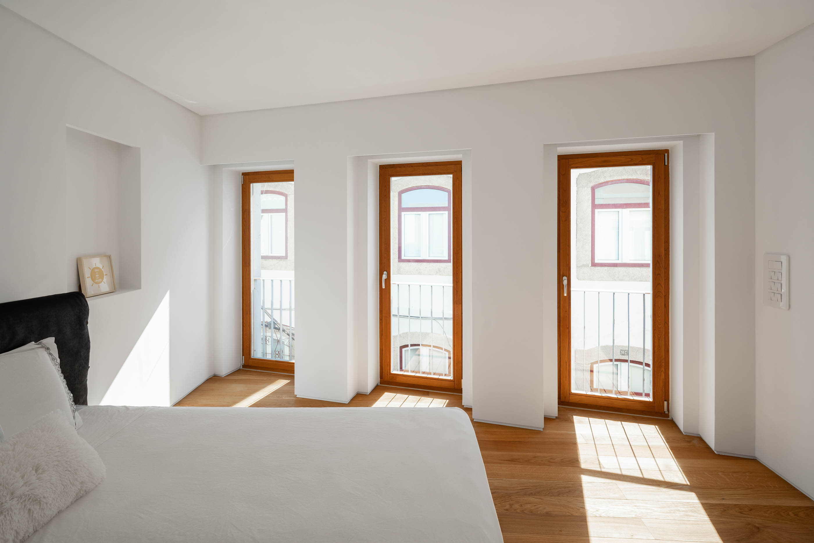 Quarto com cama branca, janelas de madeira e chão de madeira clara. — Casa GD arquitetura Esquissos Arquitectura em Portugal