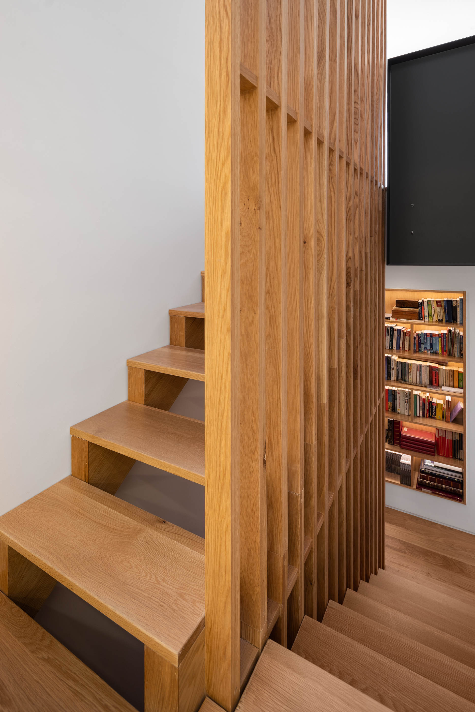 Escada de madeira clara com guarda em ripas e estante iluminada ao fundo. — Casa GD arquitetura Esquissos Arquitectura em Portugal