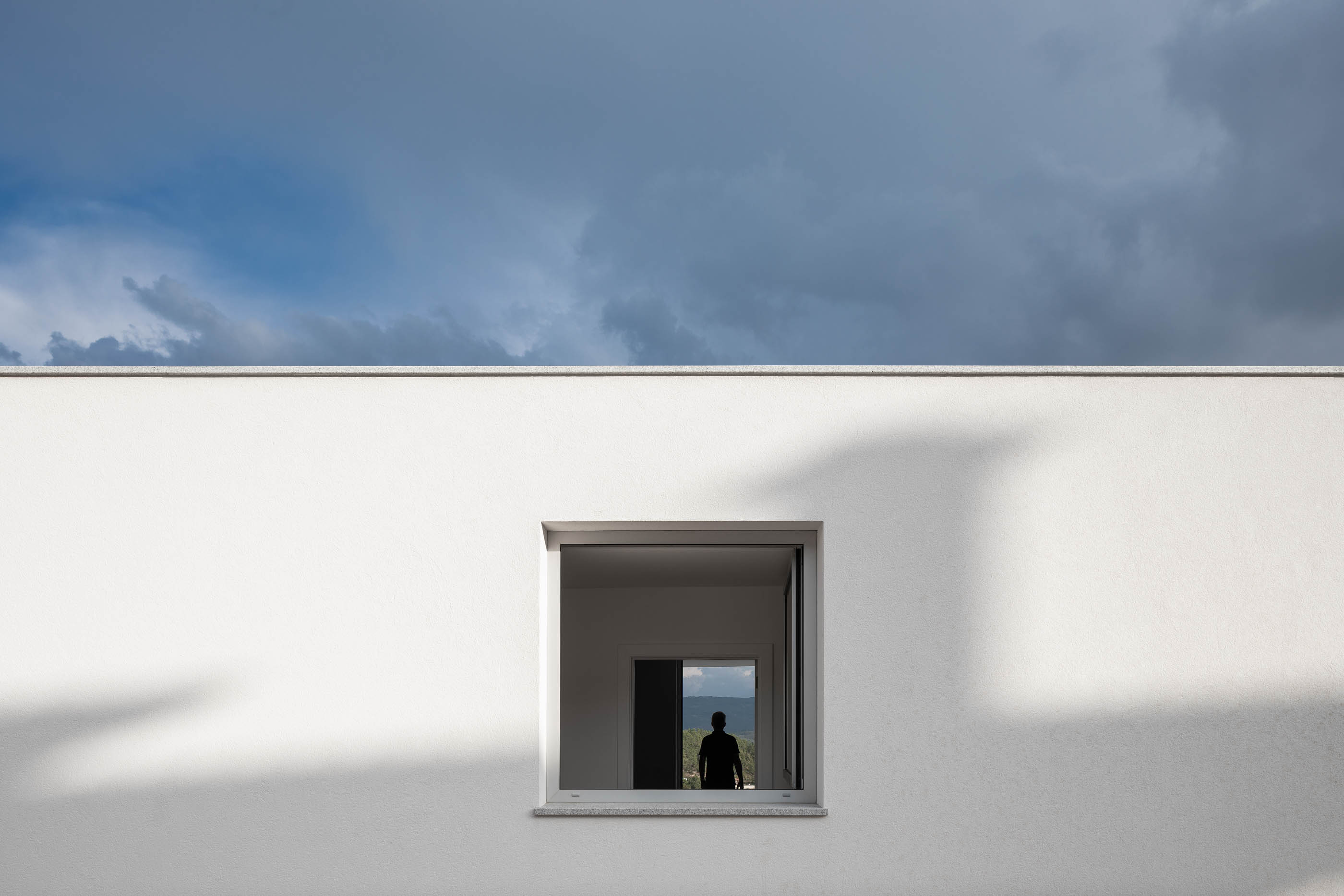 Janela quadrada numa parede branca, com silhueta de homem no interior. — Casa em Lamego arquitetura António Ildefonso Arquitectura em Portugal