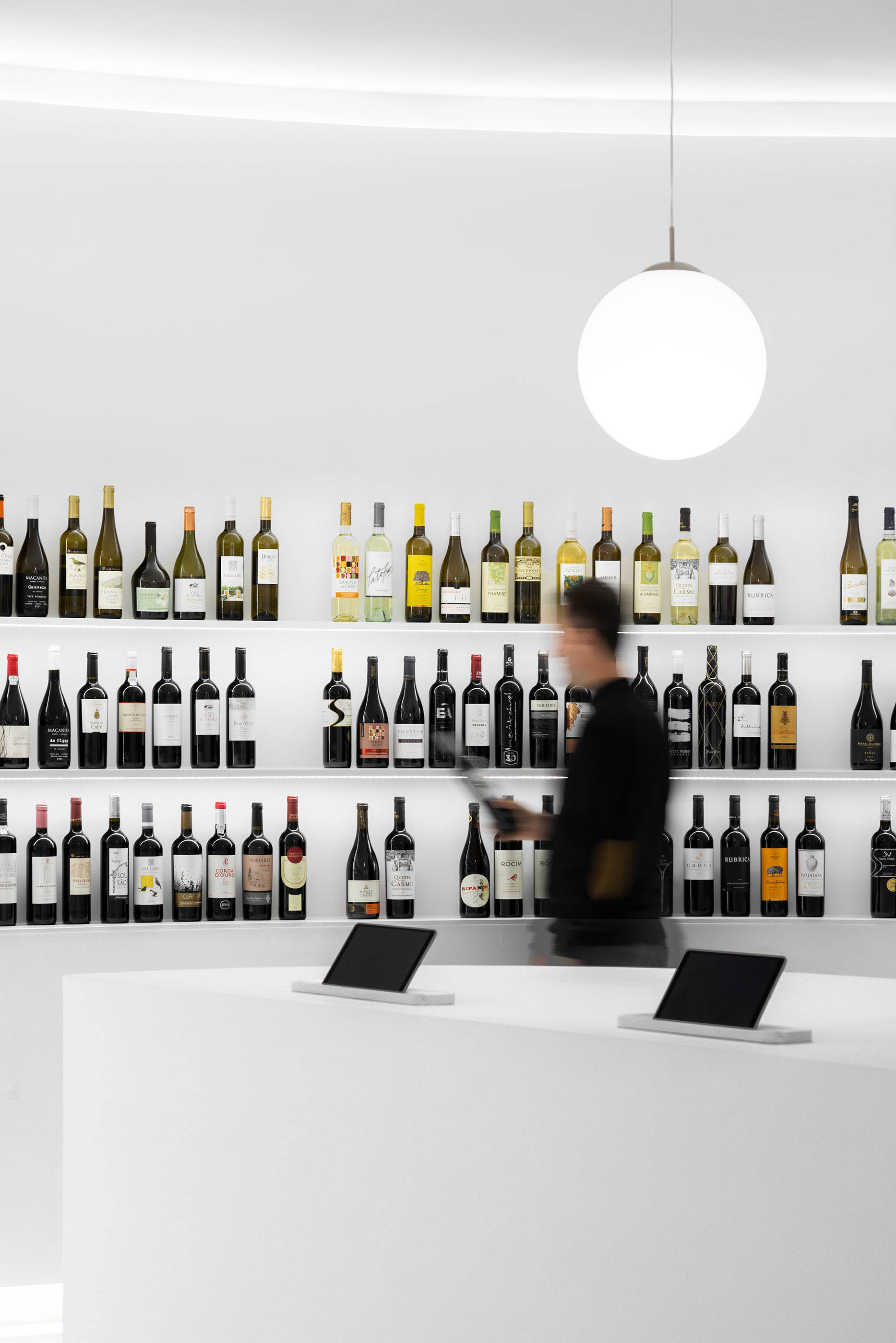 Balcão de atendimento branco com ecrãs, garrafas de vinho em prateleiras e figura humana desfocada. — Portugal Vineyards Concept Store arquitetura Porto Architects em Porto