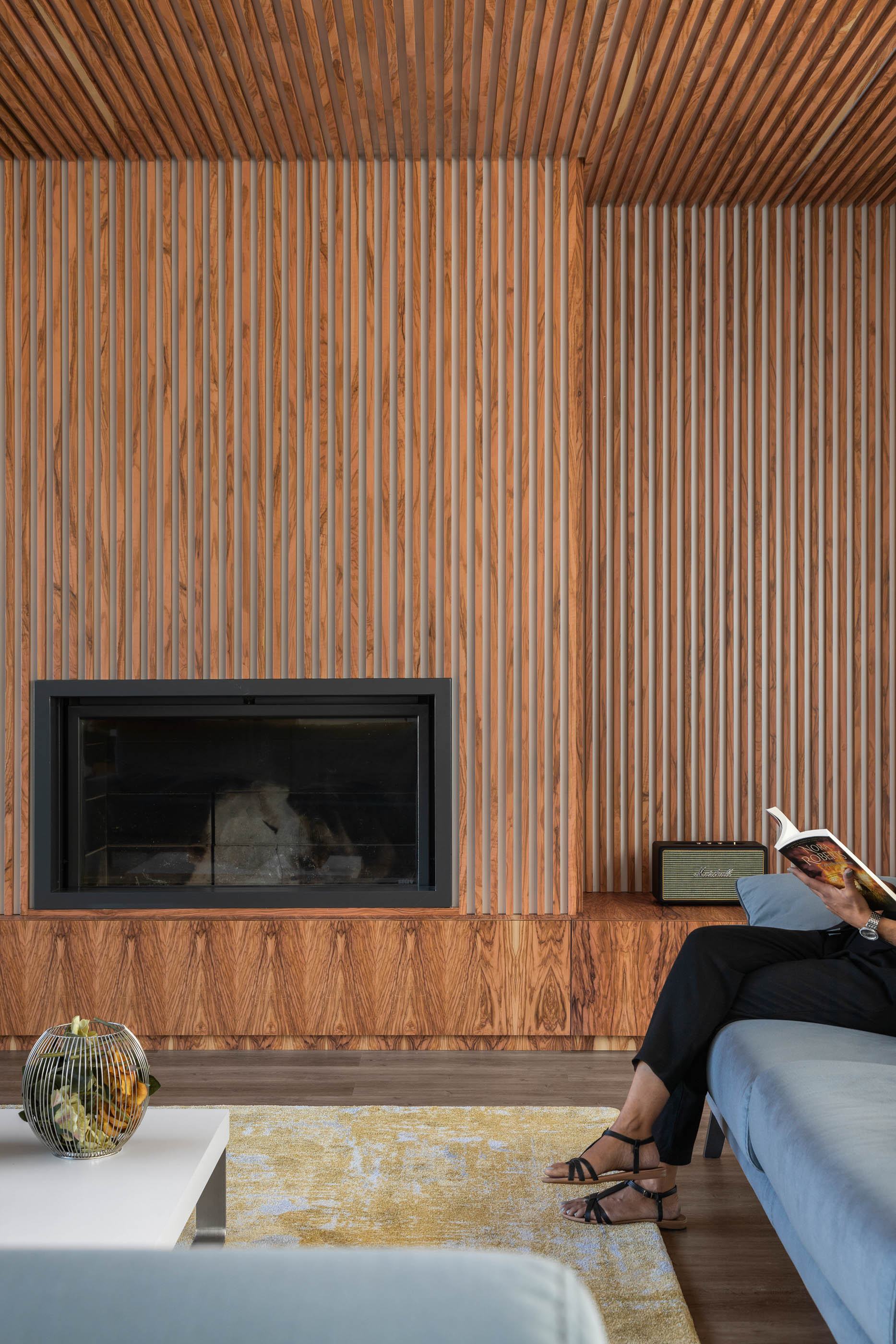 Sala de estar com lareira em madeira ripada, sofá azul e tapete amarelo, estilo moderno e acolhedor. — Reabilitação Casa J arquitetura Frari em Portugal