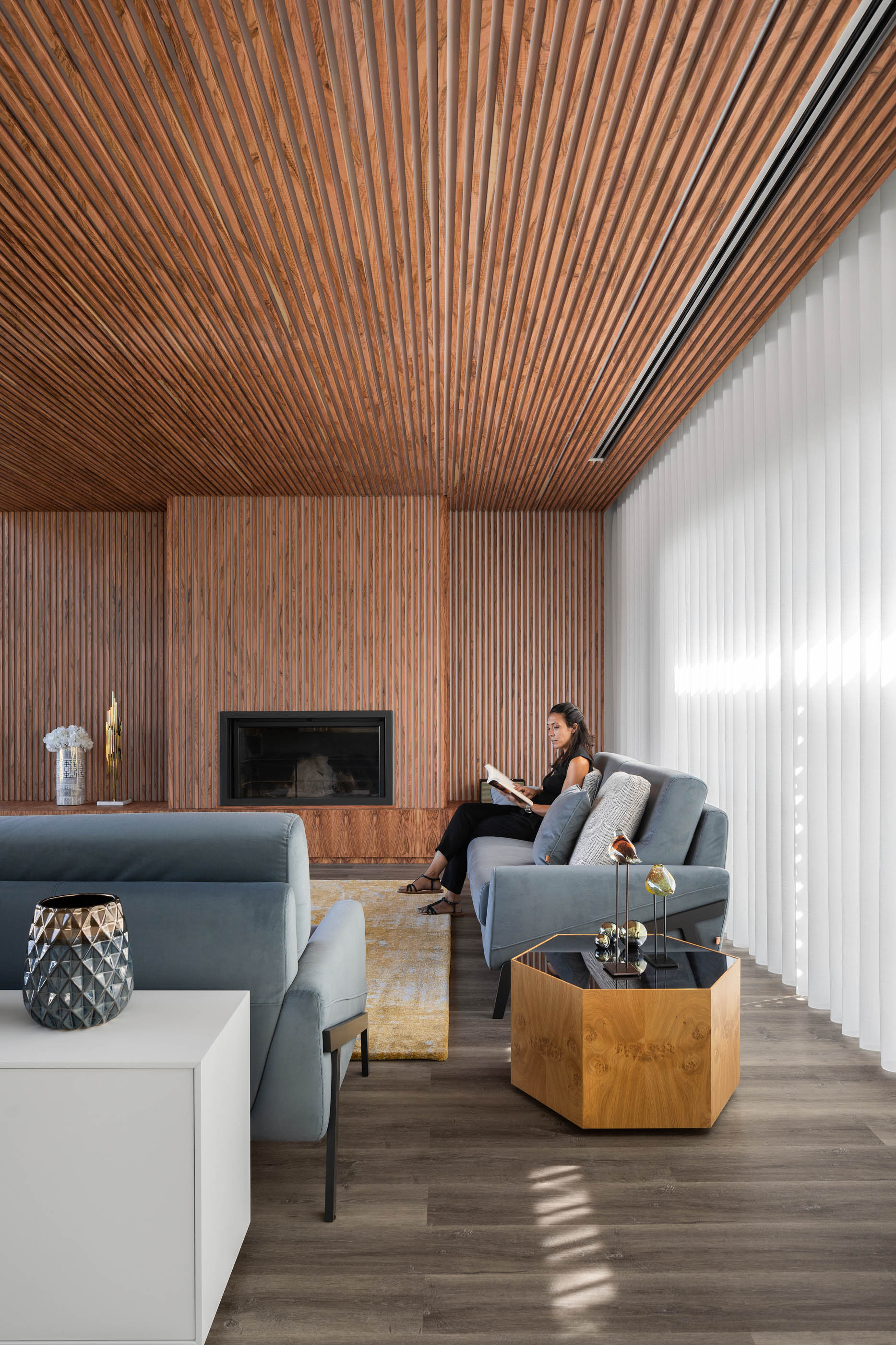 Sala de estar com teto e parede em madeira ripada, sofá azul, mesa lateral hexagonal em madeira e cortinas brancas. — Reabilitação Casa J arquitetura Frari em Portugal