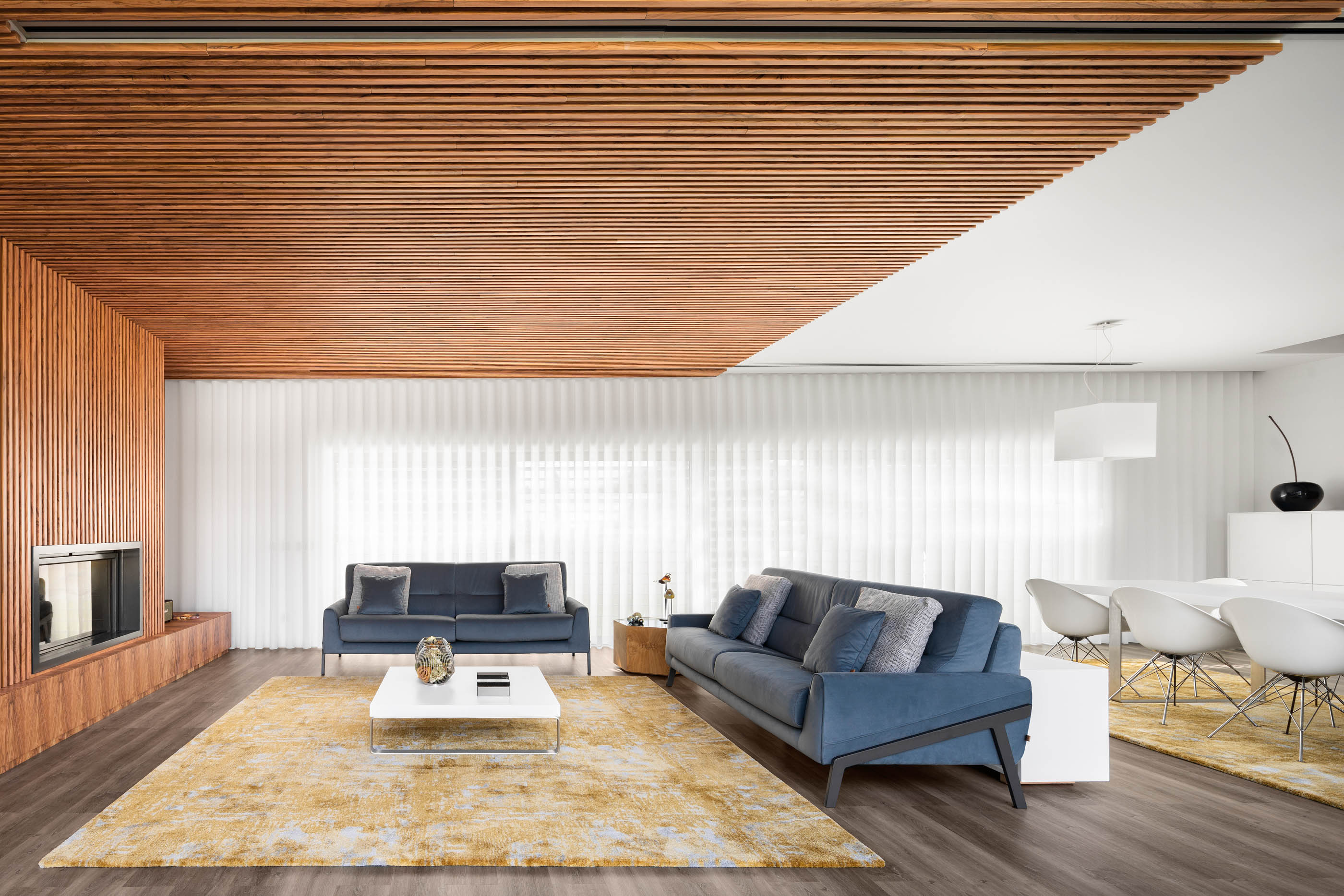 Sala ampla com teto em madeira ripada, sofás azuis, tapete amarelo e zona de refeições com cadeiras brancas. — Reabilitação Casa J arquitetura Frari em Portugal