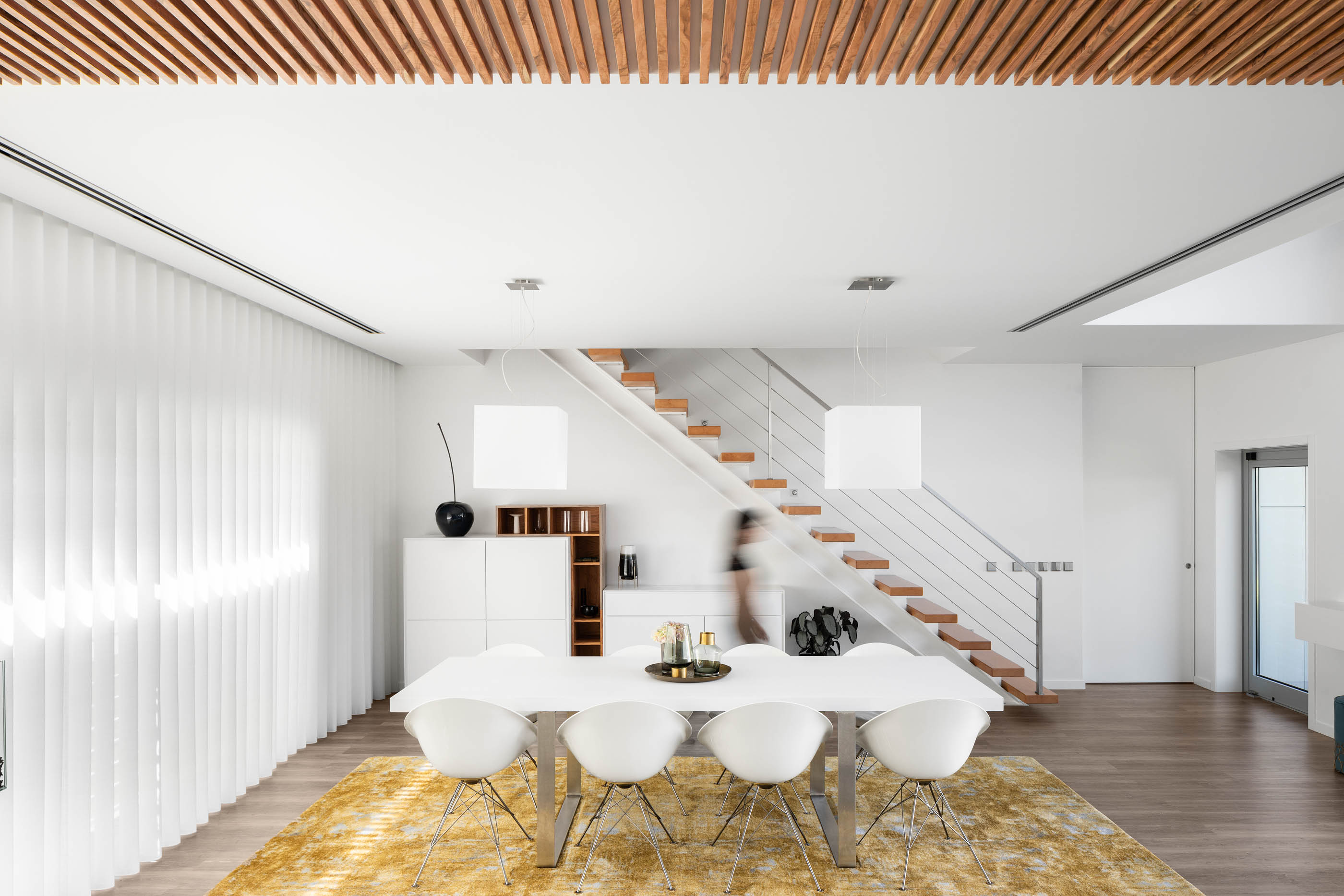 Sala de jantar com mesa e cadeiras brancas, tapete amarelo e escadas em madeira com estrutura metálica. — Reabilitação Casa J arquitetura Frari em Portugal