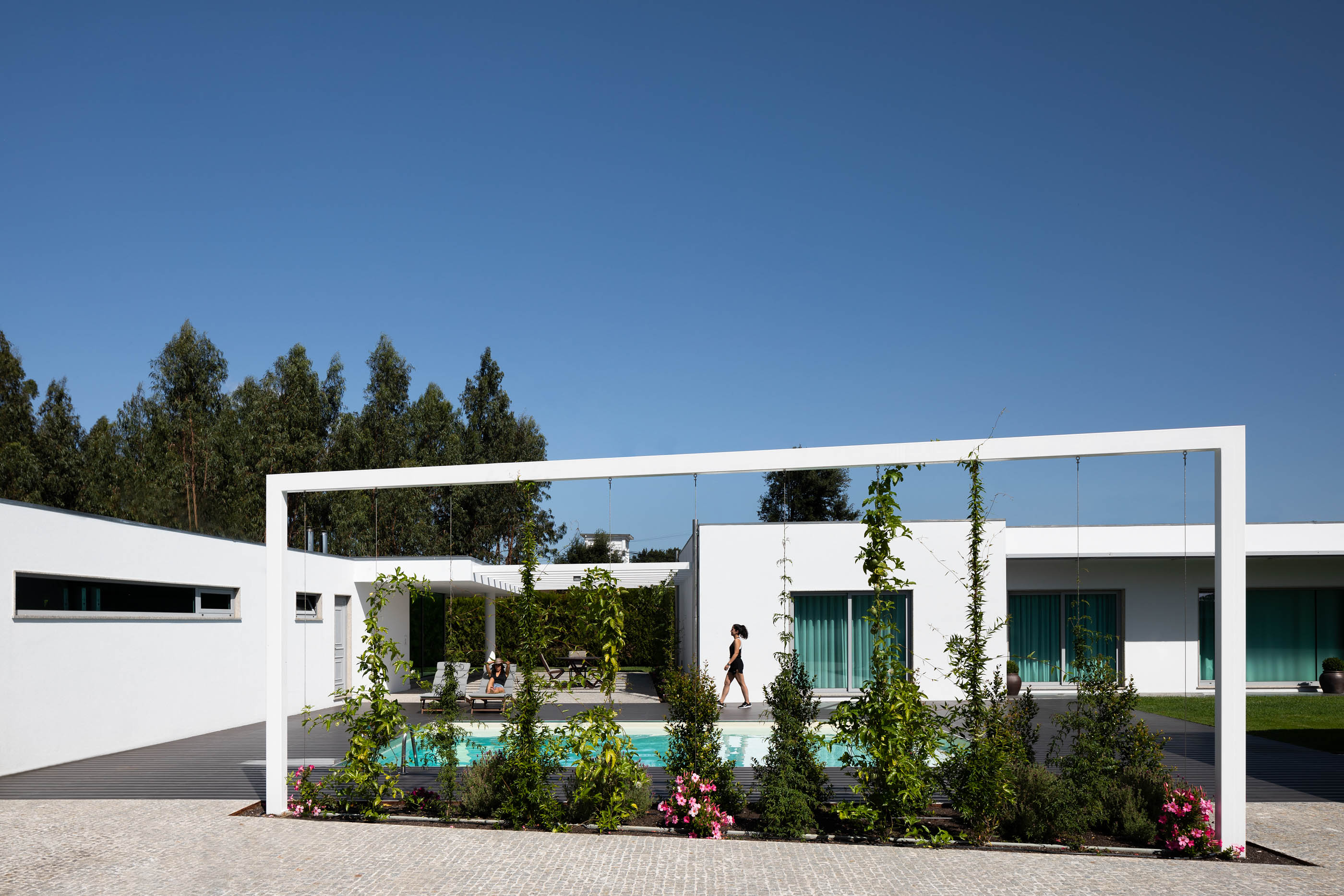 Exterior da casa com piscina, pérgola branca com plantas e relvado, num dia ensolarado. — Reabilitação Casa J arquitetura Frari em Portugal