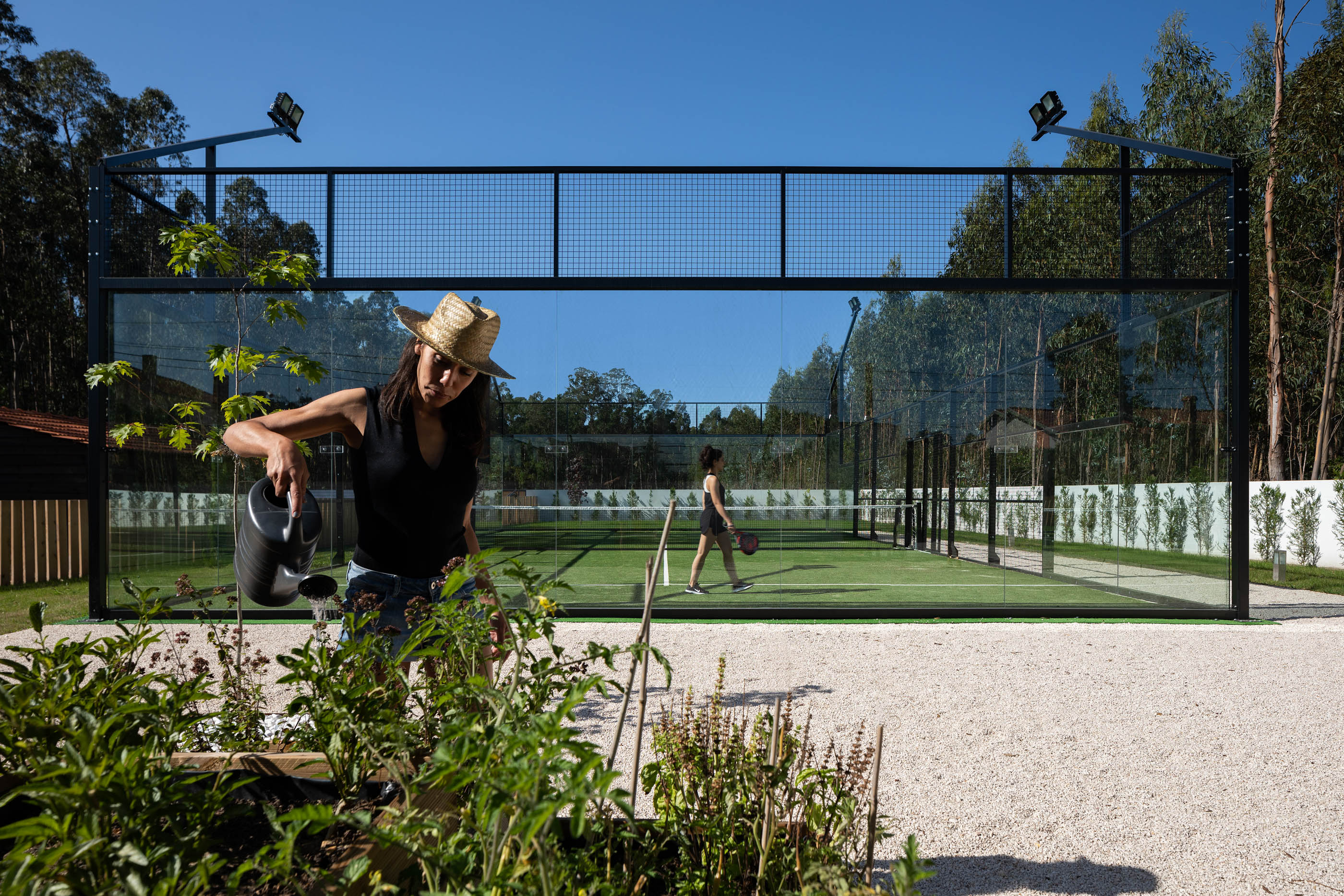 Campo de padel com estrutura metálica e paredes de vidro, com vegetação e uma pessoa a regar plantas. — Reabilitação Casa J arquitetura Frari em Portugal