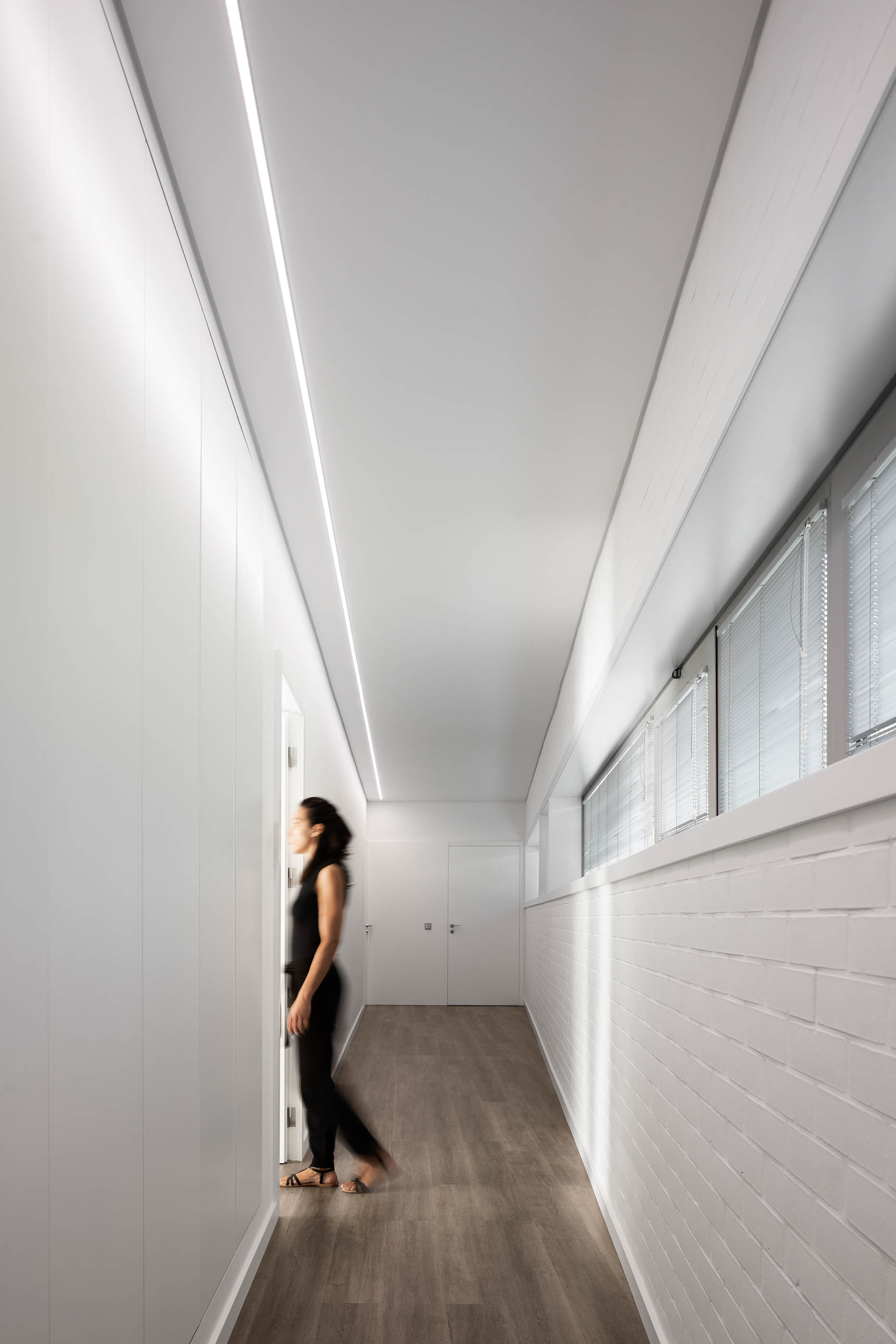 Corredor minimalista com paredes brancas, iluminação linear e pavimento em madeira, com uma pessoa a caminhar. — Reabilitação Casa J arquitetura Frari em Portugal