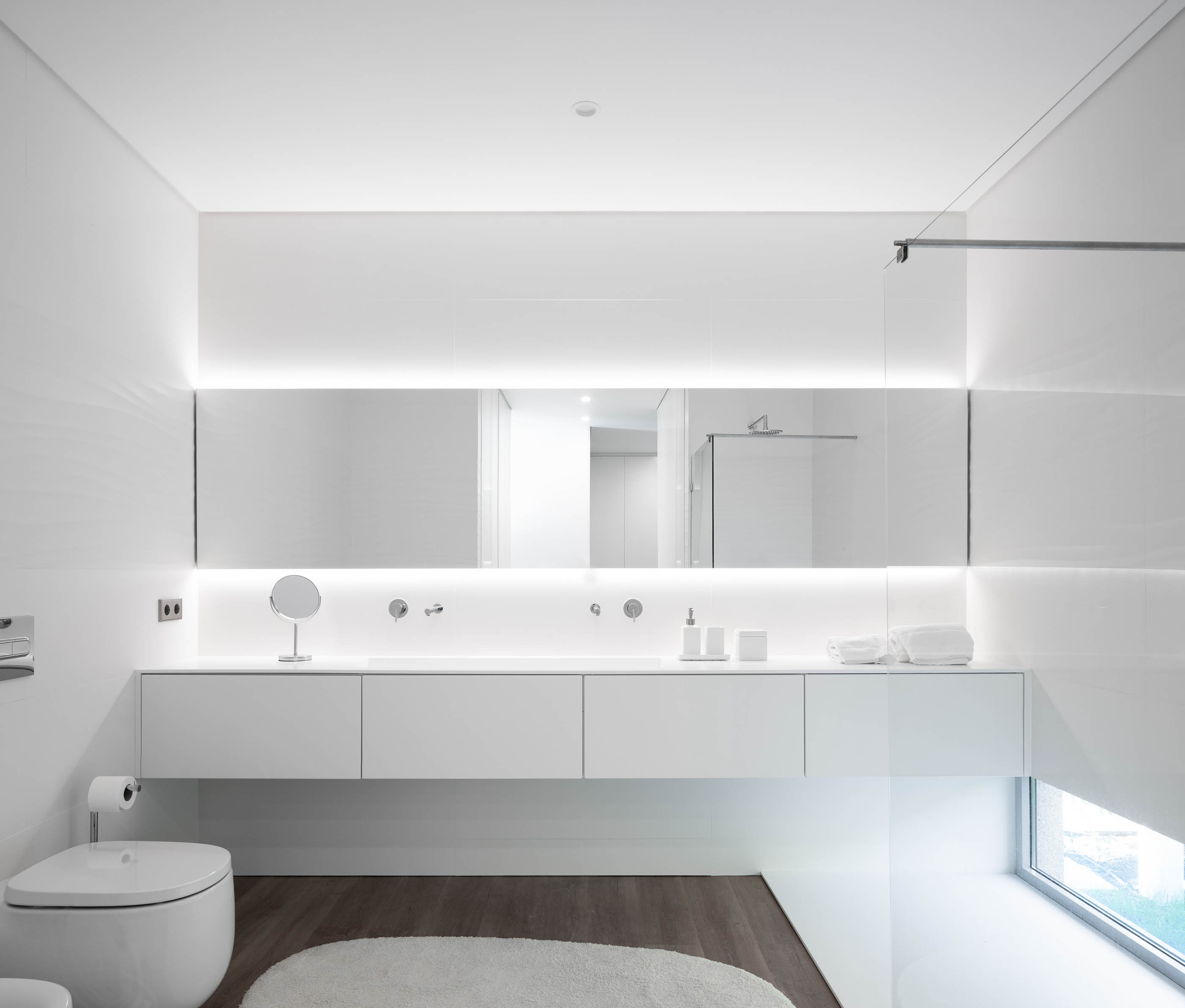 Casa de banho minimalista com lavatório suspenso branco, espelho amplo com iluminação e pavimento em madeira. — Reabilitação Casa J arquitetura Frari em Portugal
