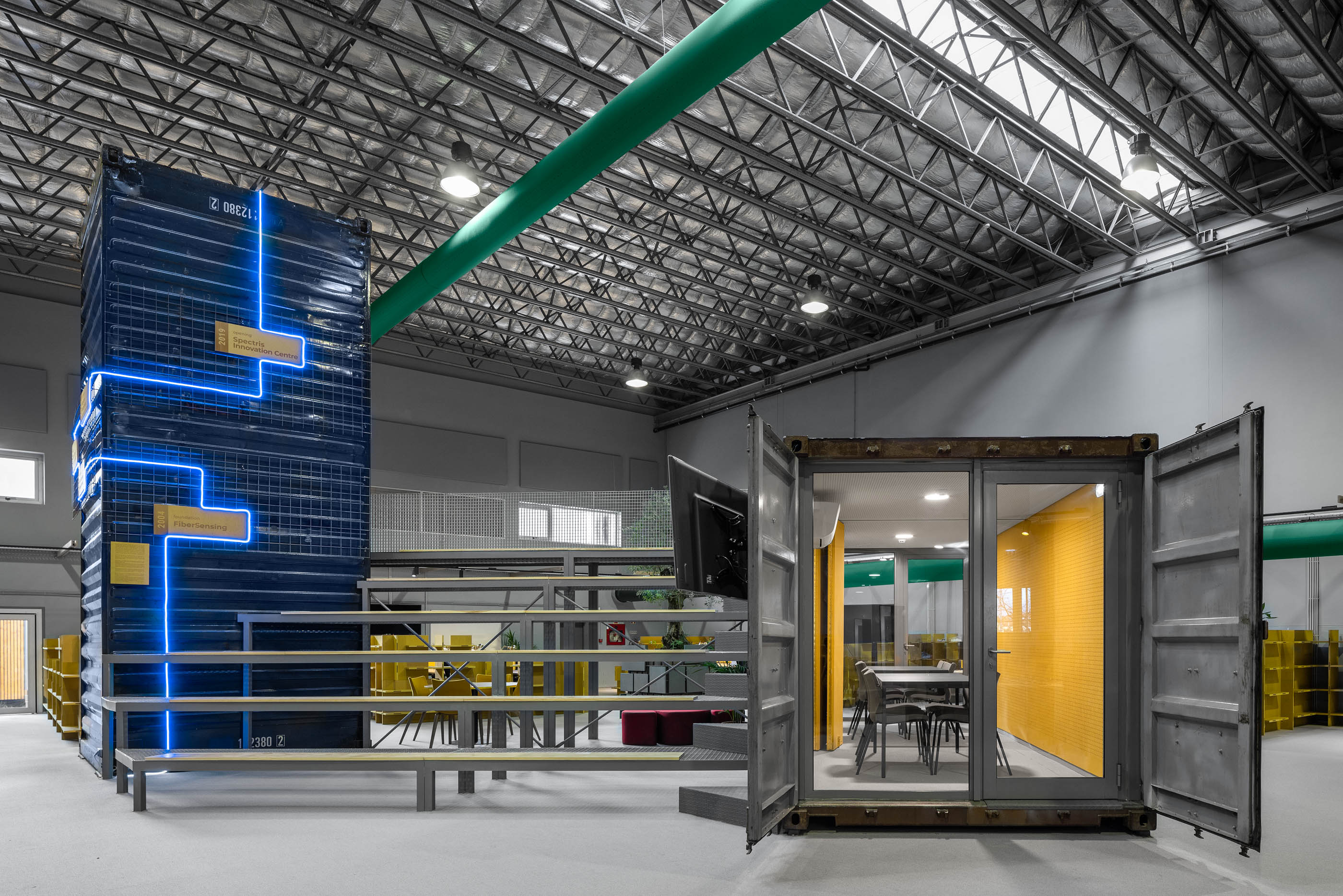 Espaço amplo: Estruturas metálicas, contentores marítimos transformados em escritórios, iluminação industrial e mobiliário colorido. — HBK Spectris Innovation Centre arquitetura Studium Creative Studio em Porto