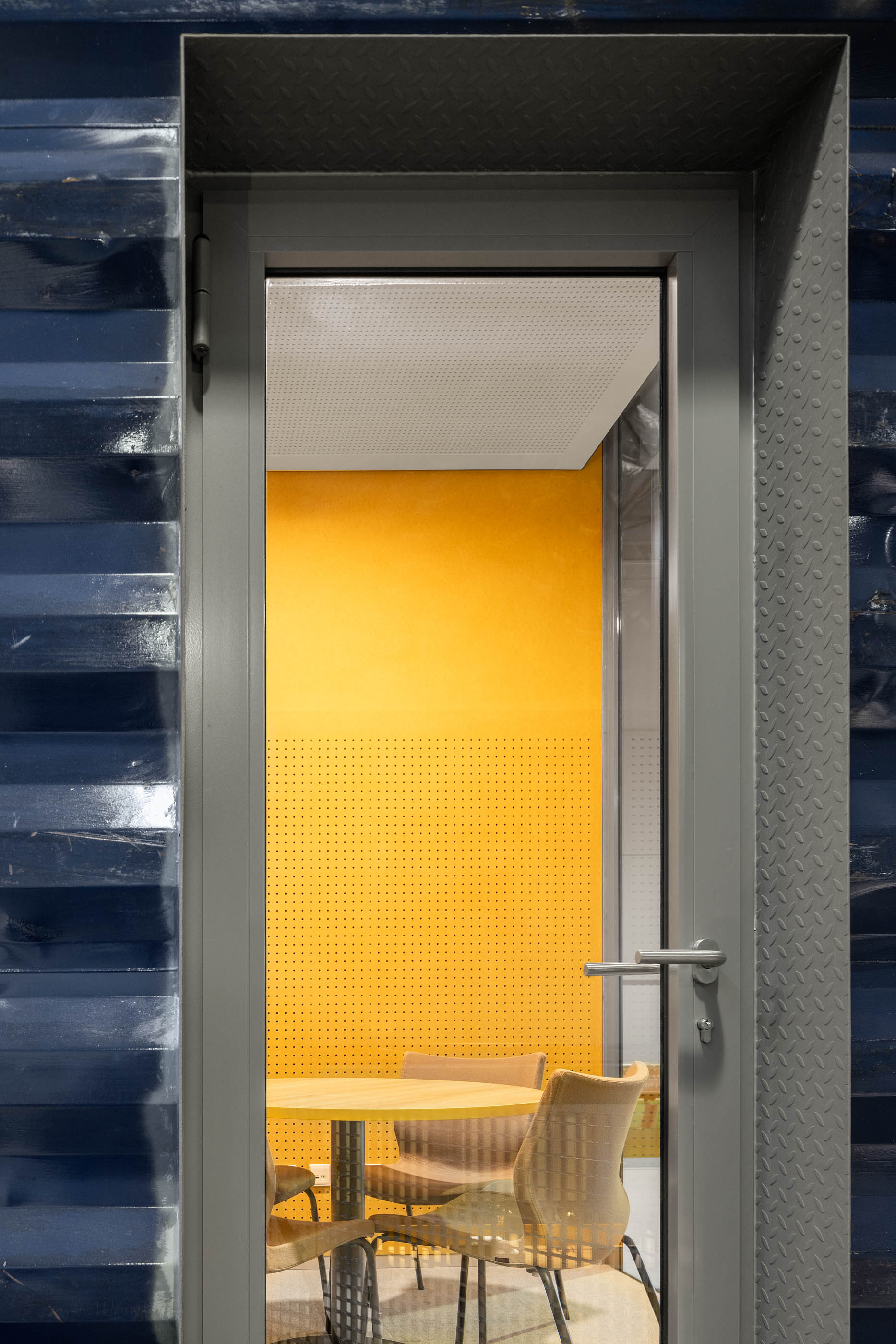 Detalhe: Porta metálica cinzenta de um contentor, vista para interior com parede amarela perfurada e mobiliário de escritório. — HBK Spectris Innovation Centre arquitetura Studium Creative Studio em Porto