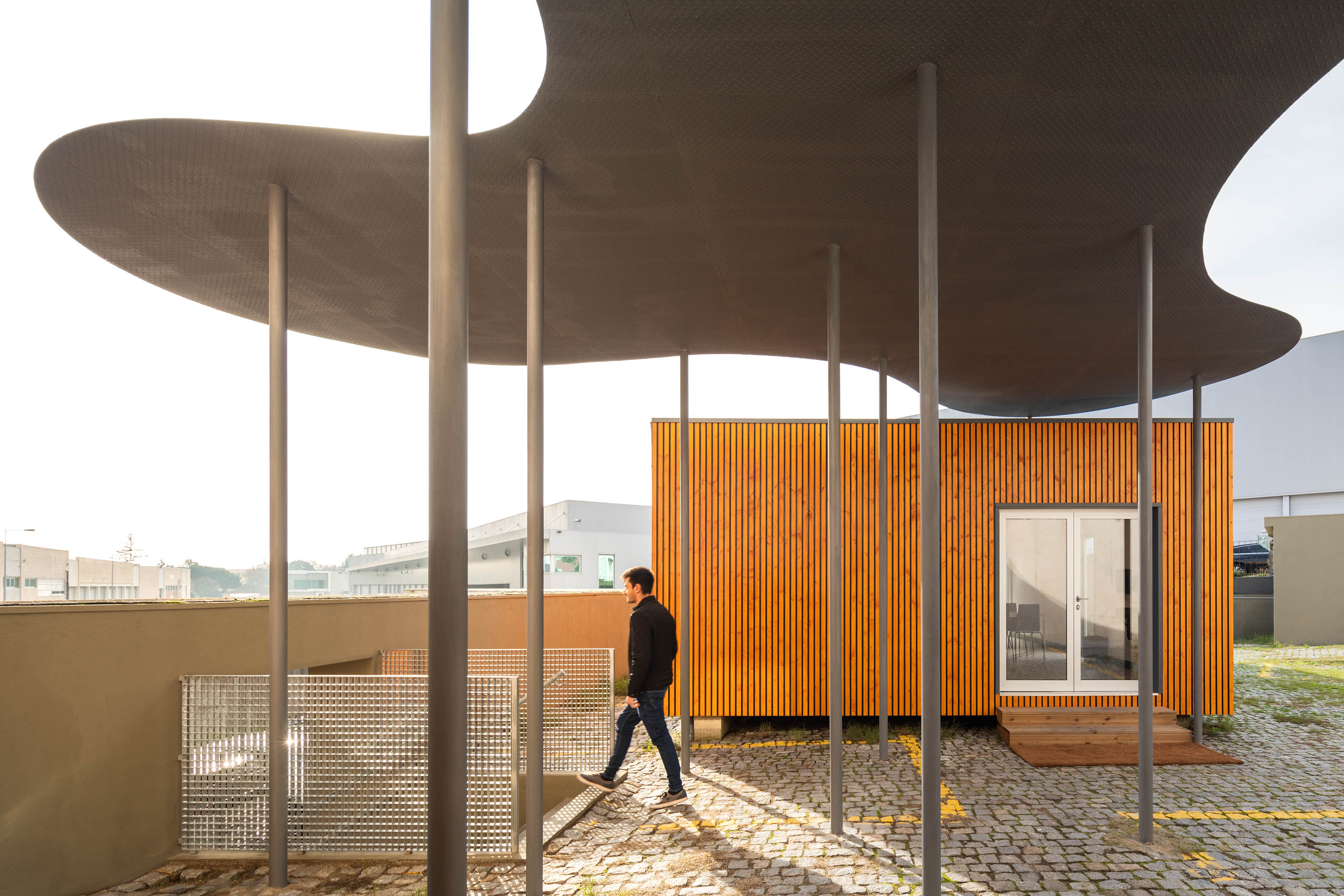Exterior: Estrutura metálica com cobertura orgânica, fachada em madeira e pavimentação em calçada. — HBK Spectris Innovation Centre arquitetura Studium Creative Studio em Porto