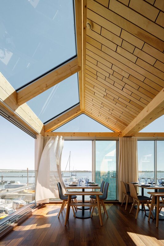 Divisão interior com mesas postas, vista para a água, teto em madeira clara com vigas expostas. — Restaurante Clube de Vela da Costa Nova arquitetura Ferreira Arquitetos em Ílhavo