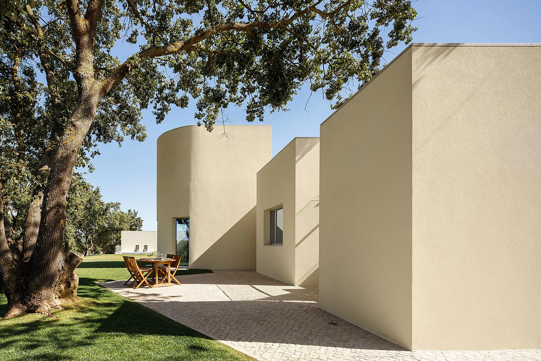 Fachada da casa com volumes geométricos, revestimento bege e calçada portuguesa. Árvore frondosa e mobiliário de exterior em madeira. — Casa em Santarém arquitetura dp arquitectos em Santarém