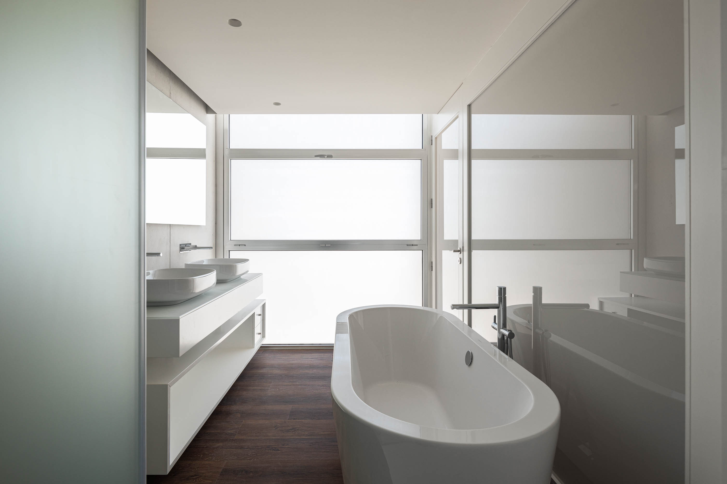 Casa de banho: Banheira oval branca, lavatórios duplos sobre móvel branco, pavimento em madeira escura. — Edifício ORIZZONT arquitetura RVDM - Ricardo Vieira de Melo, arquitectos em Aveiro