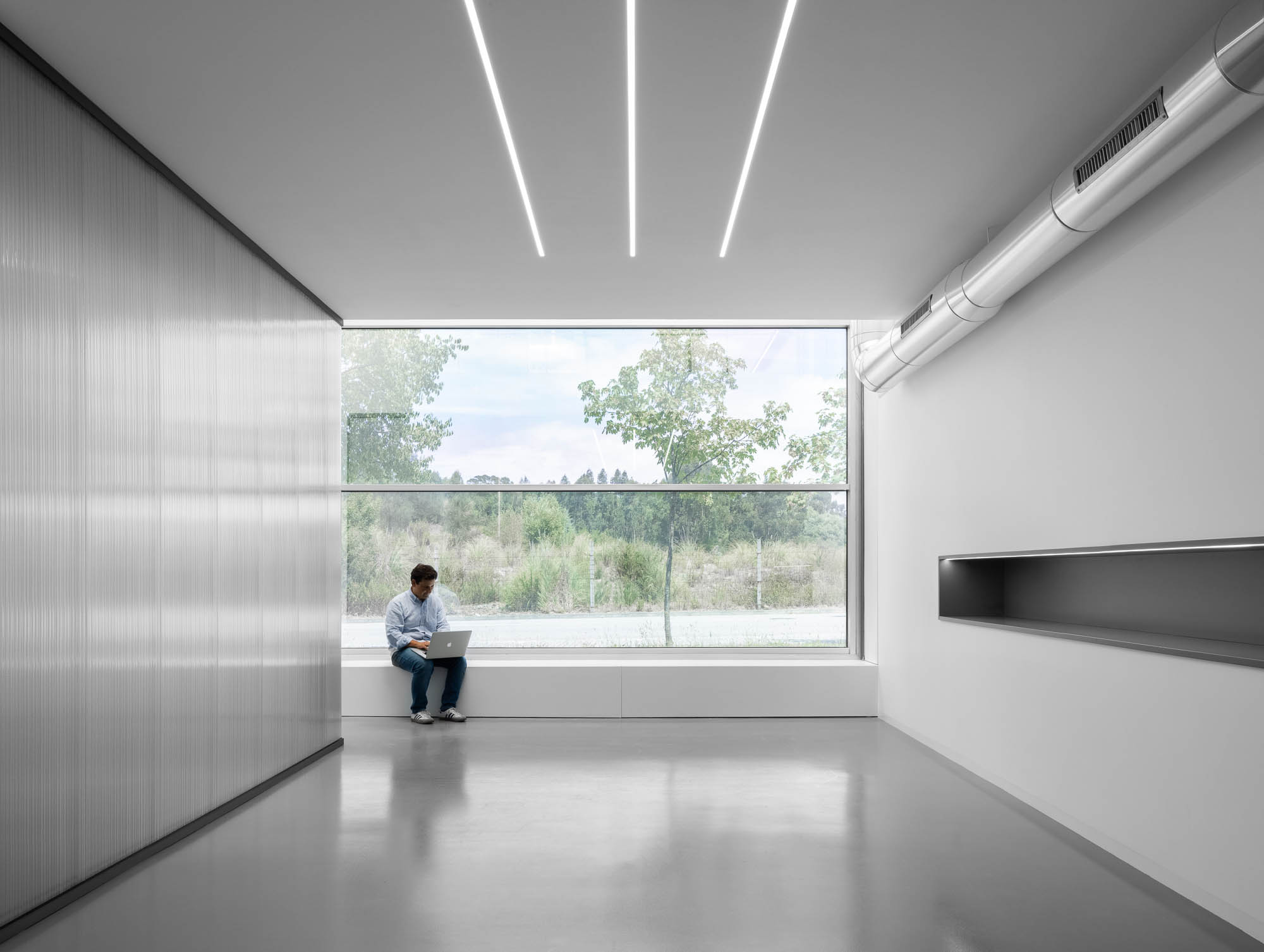 Escritório minimalista com chão cinzento polido, parede revestida a painéis metálicos e grande janela com vista para o exterior. — Escritórios MA arquitetura éOp – arquitectura e design em Maia