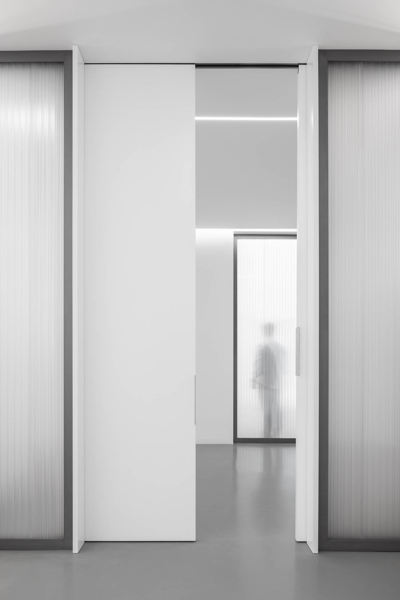 Perspectiva de corredor com portas deslizantes brancas abertas, paredes em policarbonato e silhueta de pessoa ao fundo. — Escritórios MA arquitetura éOp – arquitectura e design em Maia
