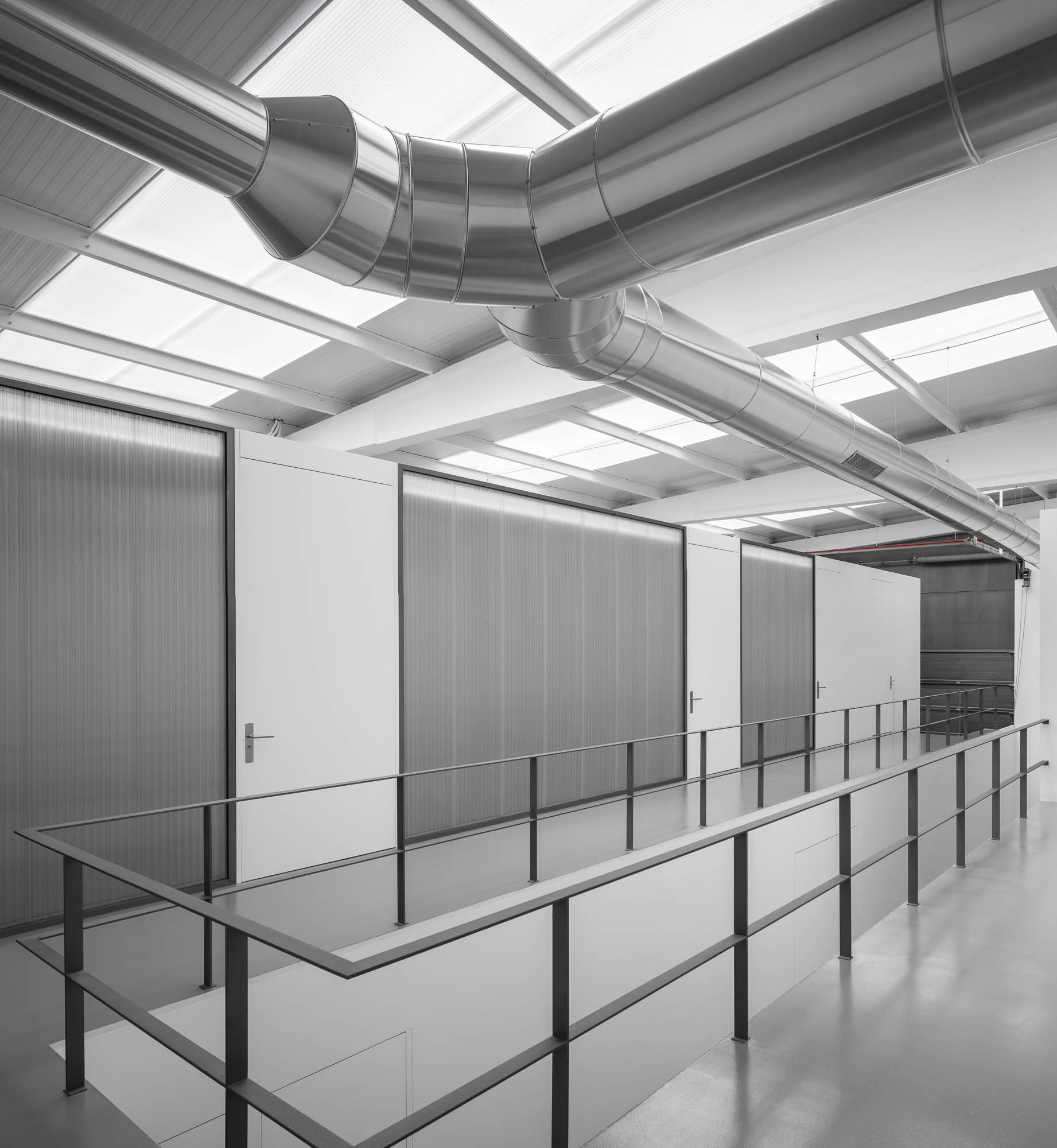 Corredor elevado com guarda-corpo metálico, paredes em policarbonato e teto com claraboias e tubagens expostas. — Escritórios MA arquitetura éOp – arquitectura e design em Maia