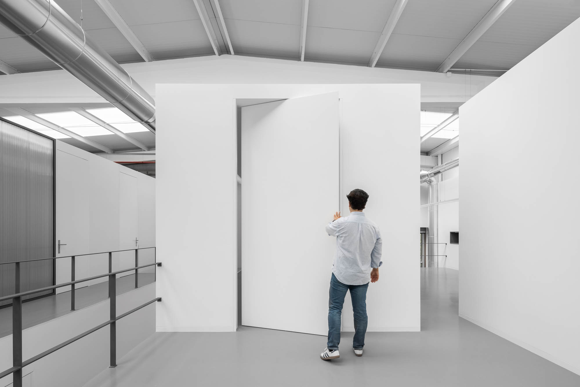 Espaço amplo com estrutura cúbica branca e porta pivotante semi-aberta, tubagens expostas no teto e guarda-corpo metálico. — Escritórios MA arquitetura éOp – arquitectura e design em Maia