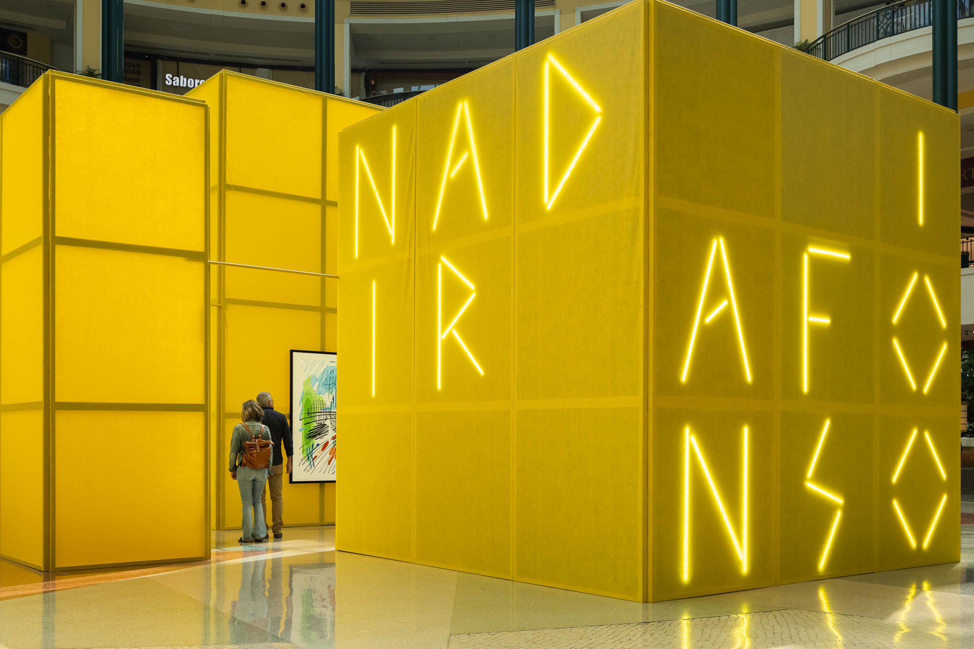 Divisão com cubos amarelos translúcidos, iluminação neon com o nome do artista, e visitantes a apreciar uma pintura. — Museu Temporário Nadir Afonso arquitetura Diogo Aguiar Studio em Lisboa
