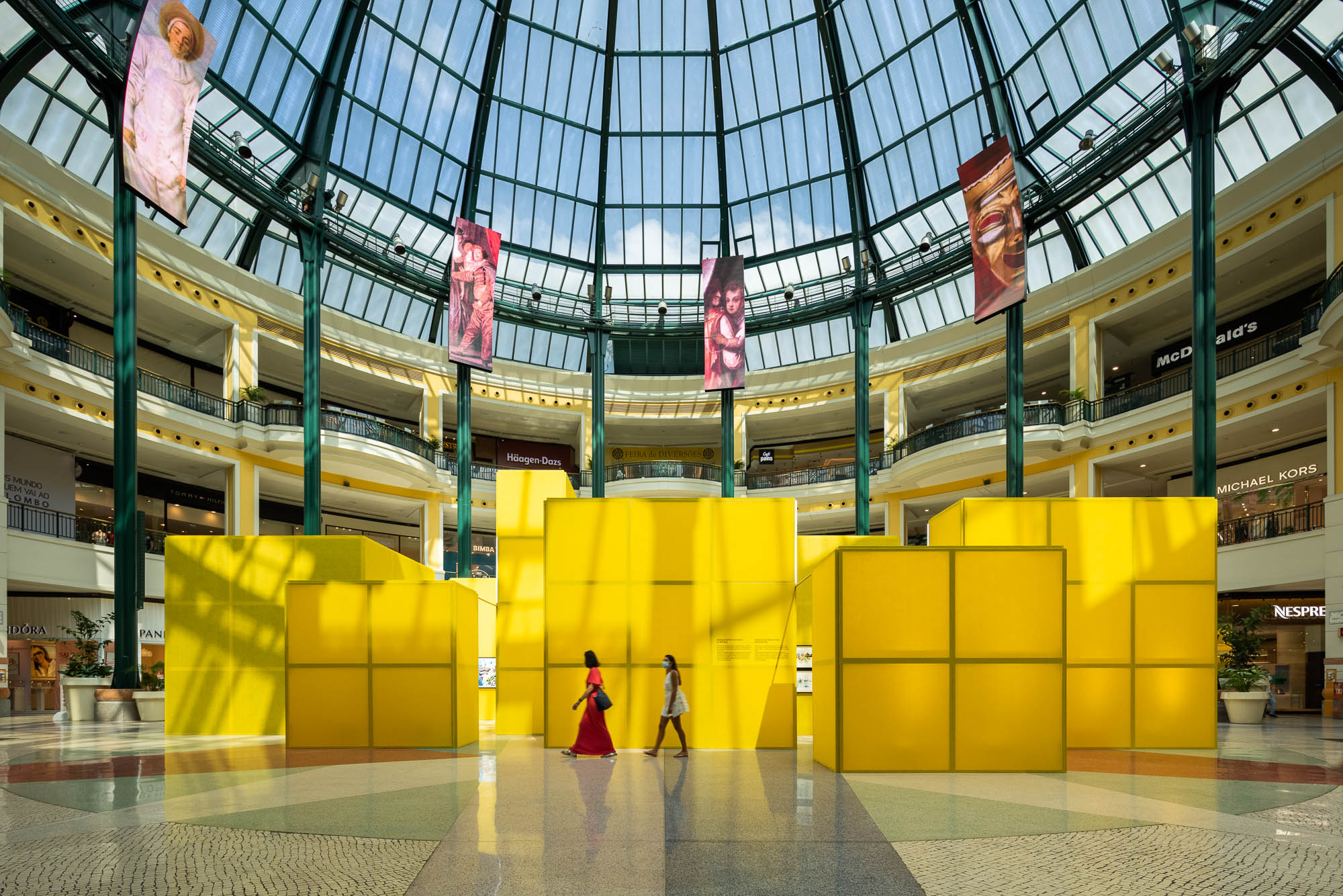 Vista geral da instalação com cubos amarelos num centro comercial, sob uma cúpula de vidro, com pessoas a circular. — Museu Temporário Nadir Afonso arquitetura Diogo Aguiar Studio em Lisboa