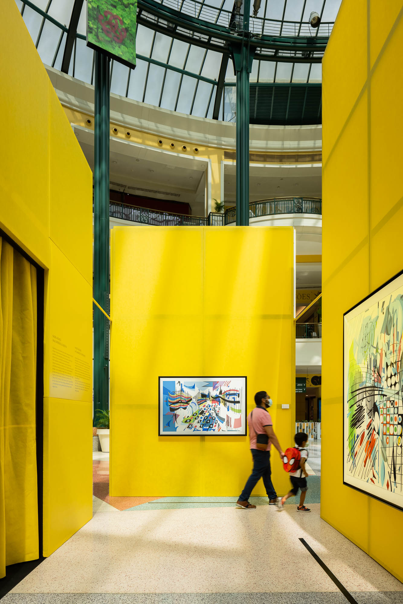 Corredor entre painéis amarelos, com obras de arte em exibição e visitantes a percorrer o espaço. — Museu Temporário Nadir Afonso arquitetura Diogo Aguiar Studio em Lisboa