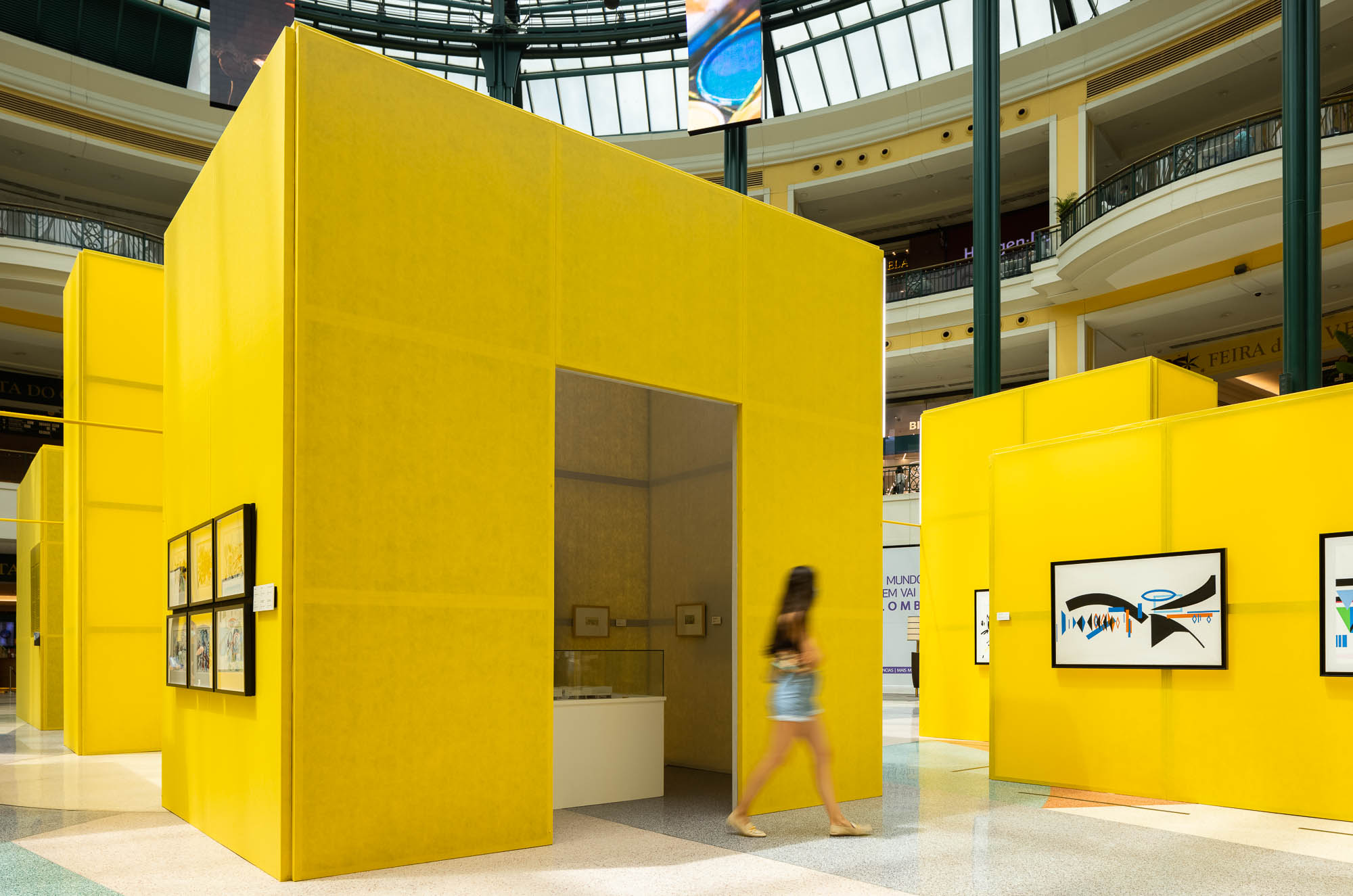 Vista geral da instalação com cubos amarelos num centro comercial, com visitantes e obras de arte. — Museu Temporário Nadir Afonso arquitetura Diogo Aguiar Studio em Lisboa