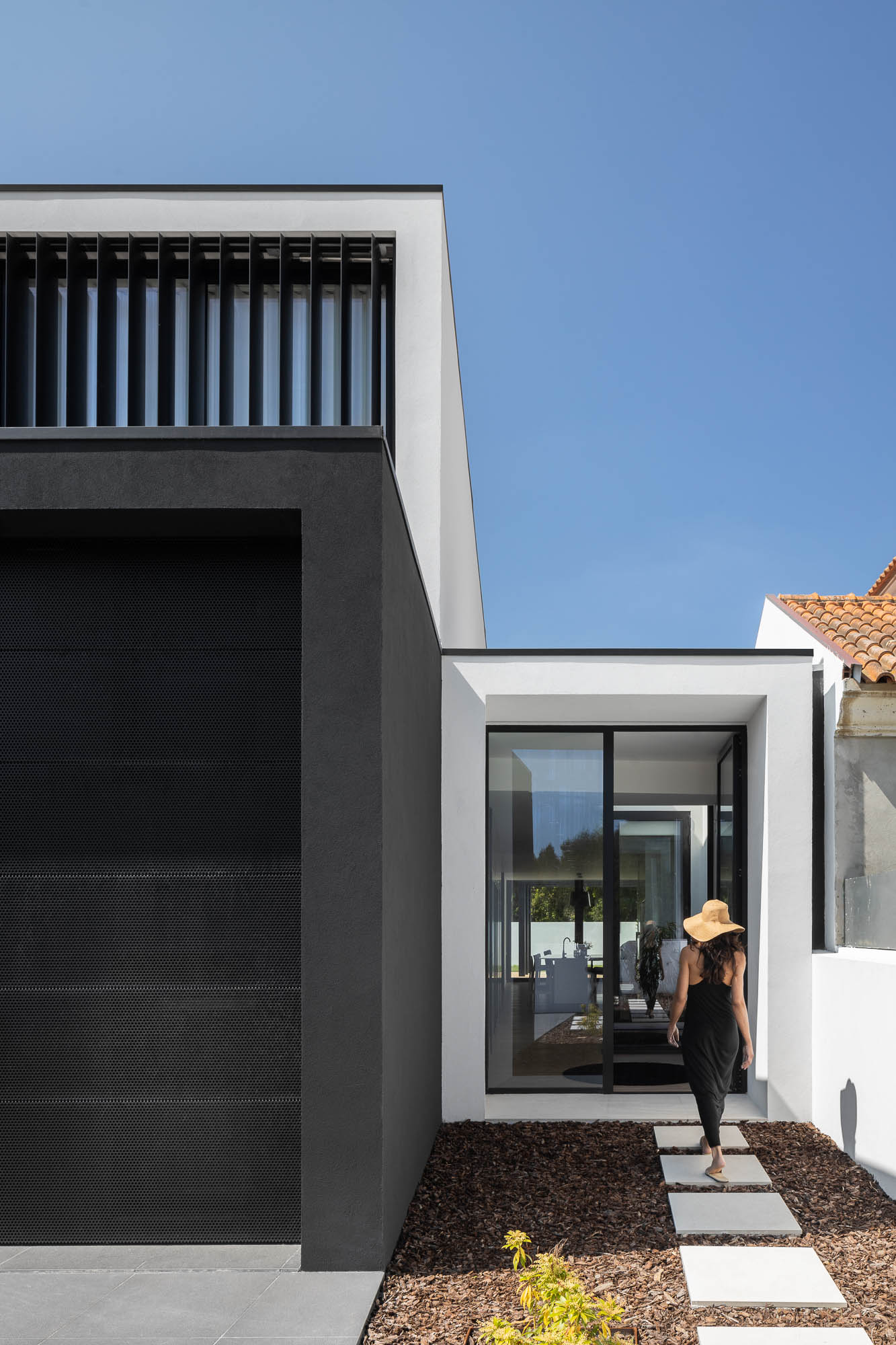 Entrada da casa com mulher a caminhar sobre passadeira de pedra, fachada branca e garagem preta. — Casa Diagonal arquitetura Frari em Aveiro