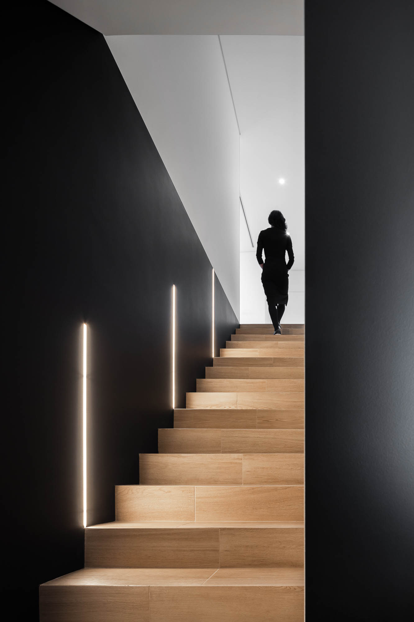 Escada em madeira com iluminação embutida vertical, paredes pretas e pessoa a subir. — Casa Diagonal arquitetura Frari em Aveiro