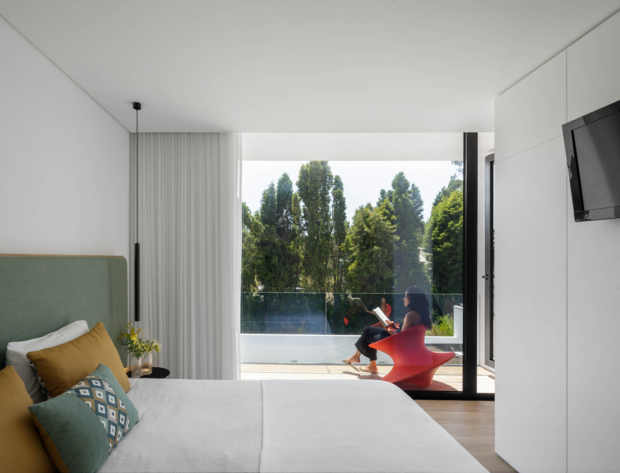Quarto com cama, varanda ampla e mulher sentada numa cadeira laranja com vista para o jardim. — Casa Diagonal arquitetura Frari em Aveiro
