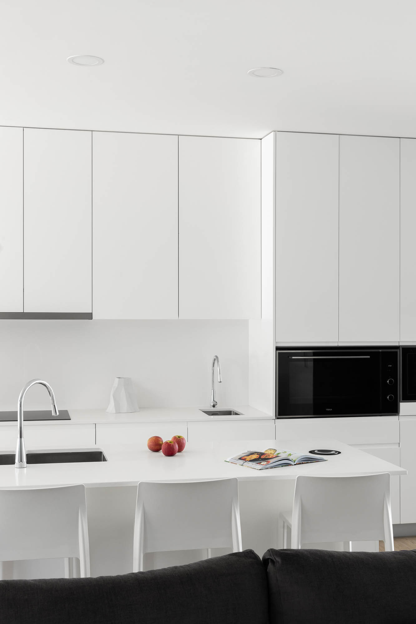 Cozinha minimalista branca com ilha central, cadeiras brancas e eletrodomésticos pretos. — Casa Diagonal arquitetura Frari em Aveiro