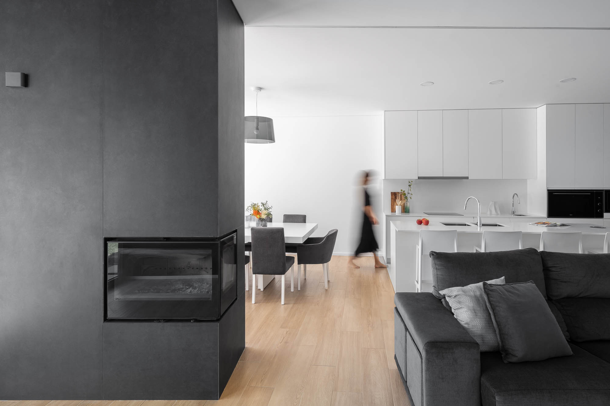 Sala de estar com lareira preta, sofá cinzento e cozinha branca ao fundo, em plano aberto. — Casa Diagonal arquitetura Frari em Aveiro