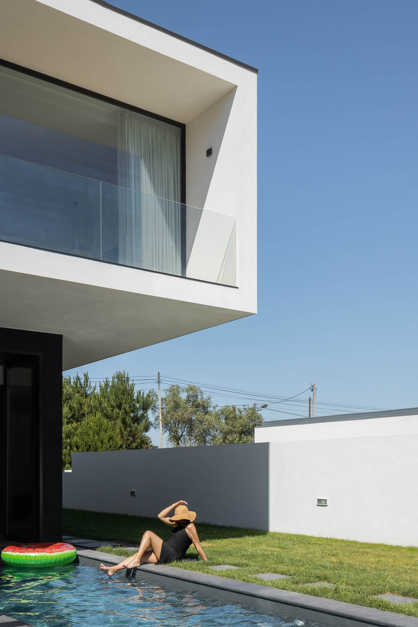 Detalhe da piscina com mulher sentada na relva, fachada branca e varanda com guarda-corpo em vidro. — Casa Diagonal arquitetura Frari em Aveiro