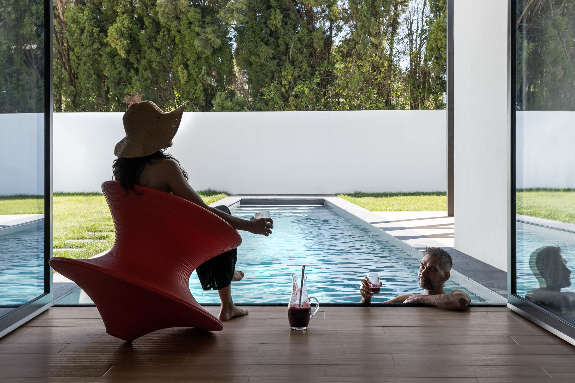 Interior com vista para a piscina, mulher sentada em cadeira laranja e homem dentro de água. — Casa Diagonal arquitetura Frari em Aveiro