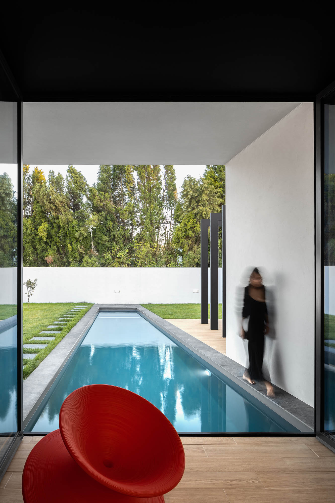 Interior com vista para a piscina, cadeira vermelha e mulher a caminhar junto à piscina. — Casa Diagonal arquitetura Frari em Aveiro